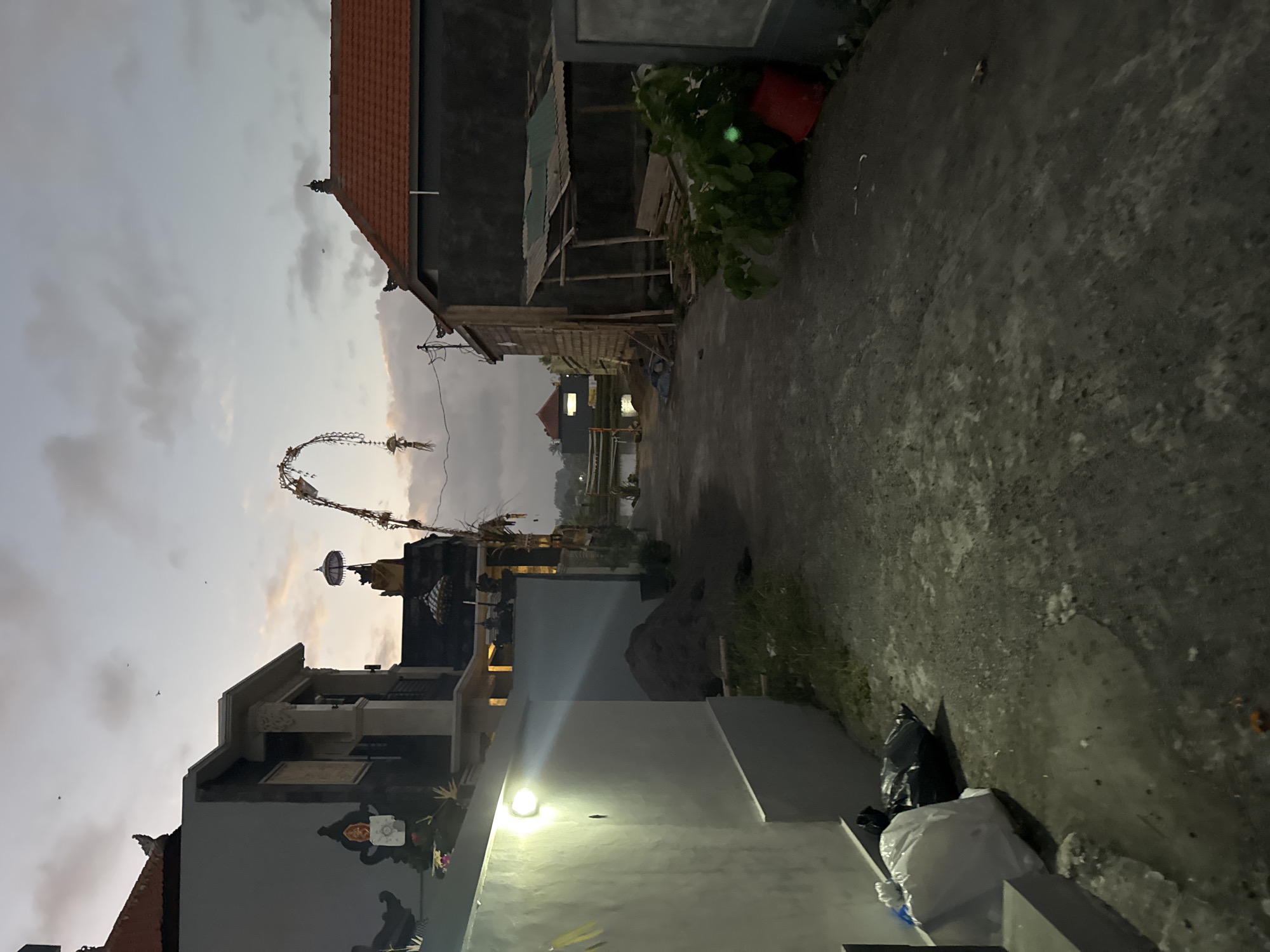 This media file depicts a street scene in Sukawati, Indonesia, during twilight. The image is vertically oriented, with the sky and buildings appearing on the left and the ground on the right. In the foreground, there's a modern building with a light illuminating the facade, which is painted gray and adorned with artwork. To the right, a paved pathway leads into the distance, bordered by grass and some piled-up trash bags.
Further into the scene, several structures are visible. There are houses with traditional tiled roofs and darker, modern-looking walls. An archway decorated with what appears to be traditional Balinese offerings or decorations stands prominently. Behind this, more buildings and some foliage are present. In the background, a body of water, possibly a rice paddy, is visible, reflecting the dim light of the sky.
The time of day is clearly evening, indicated by the soft, diffused light and the approaching darkness. The sky is overcast with subtle hints of sunset colors. The overall atmosphere suggests a quiet moment in a residential area, possibly after a day's activities or in preparation for the night. The scene evokes a sense of everyday life in a tropical locale. No visible text is present in the image.