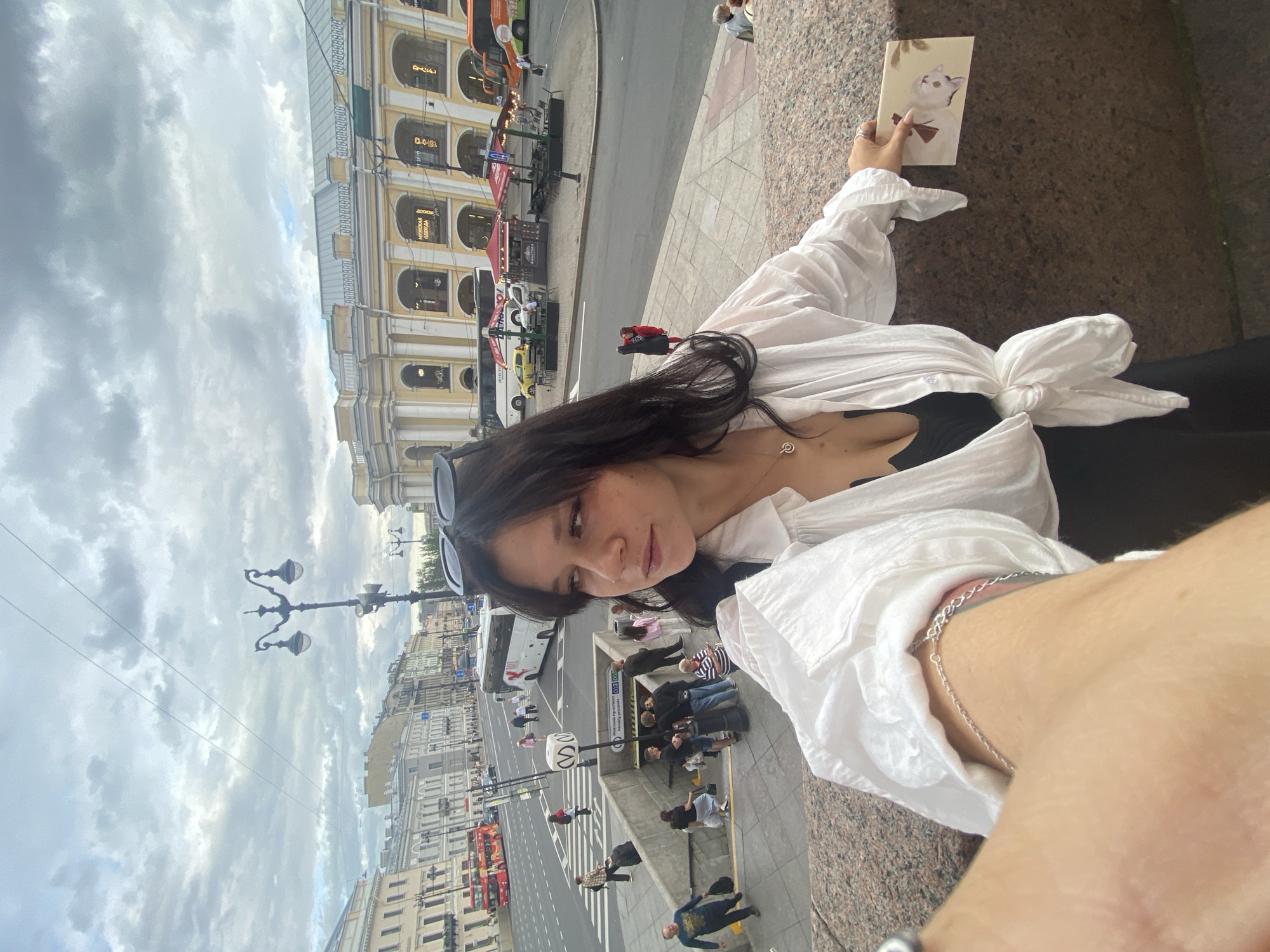 A young woman with dark hair, wearing a white open button-down shirt tied at the waist over a black top and black pants, stands on a granite staircase. She is holding a postcard with a drawing of a white cat with a red bow. She is looking to her left, away from the camera, with sunglasses perched on her head. In the background, a busy street scene in Saint Petersburg, Russia, is visible. The street features a metro station entrance, a lamppost, several buses, including a red double-decker bus, and buildings with classical architecture, including a yellow building with arched storefronts. People are walking on the street and on the stairs. The sky is cloudy.