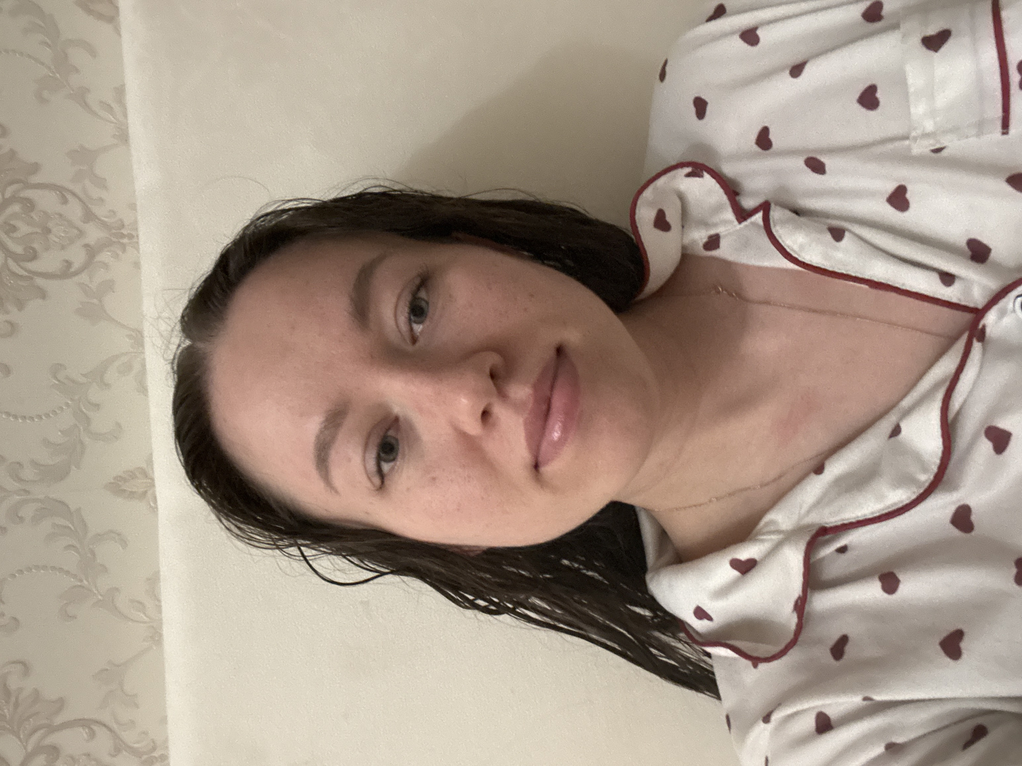 A woman with wet, dark hair lies on a light-colored surface, possibly a bed. She wears a white pajama top with a maroon heart pattern and maroon trim. A delicate gold necklace is visible on her neck. The background includes a textured, light-colored wall with a floral pattern. The location is Ферма, Russia.
