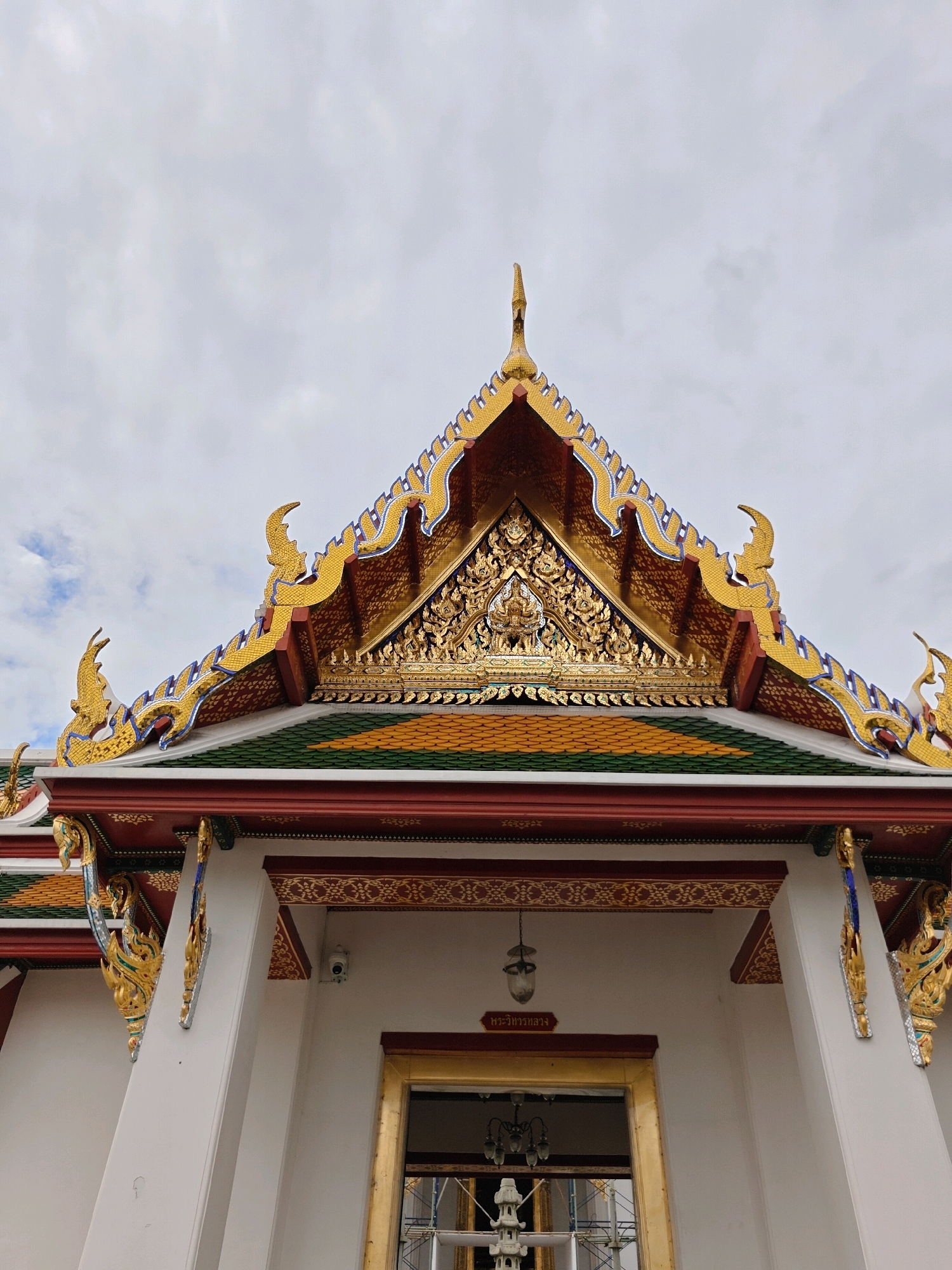 This is a low-angle shot of the ornate roof of a white building in Bangkok, Thailand. The roof features traditional Thai temple architecture with intricate gold detailing, vibrant green and yellow tiles, and decorative eaves. The sky above is overcast with clouds. The building's facade is white, with decorative elements under the eaves. A security camera is visible on the upper part of the wall. There is also a sign with Thai script above the doorway, which appears to be undergoing renovation or construction, with scaffolding visible inside. The overall impression is one of grandeur and cultural heritage.