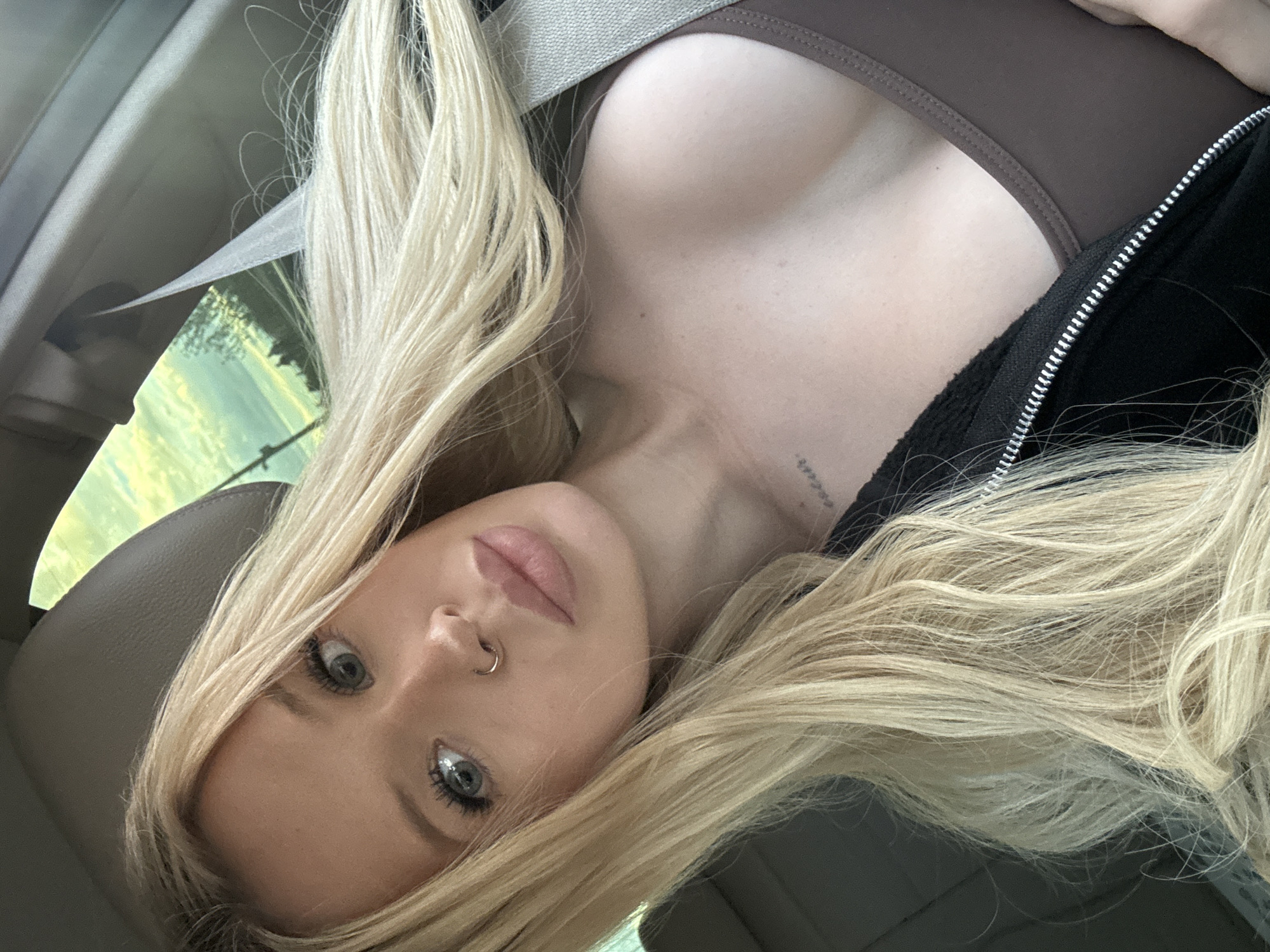 In this overhead, close-up selfie taken from inside a car, a young woman with long blonde hair and striking blue eyes is the sole subject. She is wearing a brown sports bra and a black zip-up hoodie, with the seatbelt across her chest. Her face is tilted, looking directly at the camera with full lips and a nose ring. A faint tattoo is visible on her collarbone. The car's interior is implied by the visible seatbelt and the headrest. Outside the car window, a blurred green and yellow landscape suggests daytime, possibly late afternoon given the light. The setting is Saint Petersburg, Russia. There is no visible text in the image.