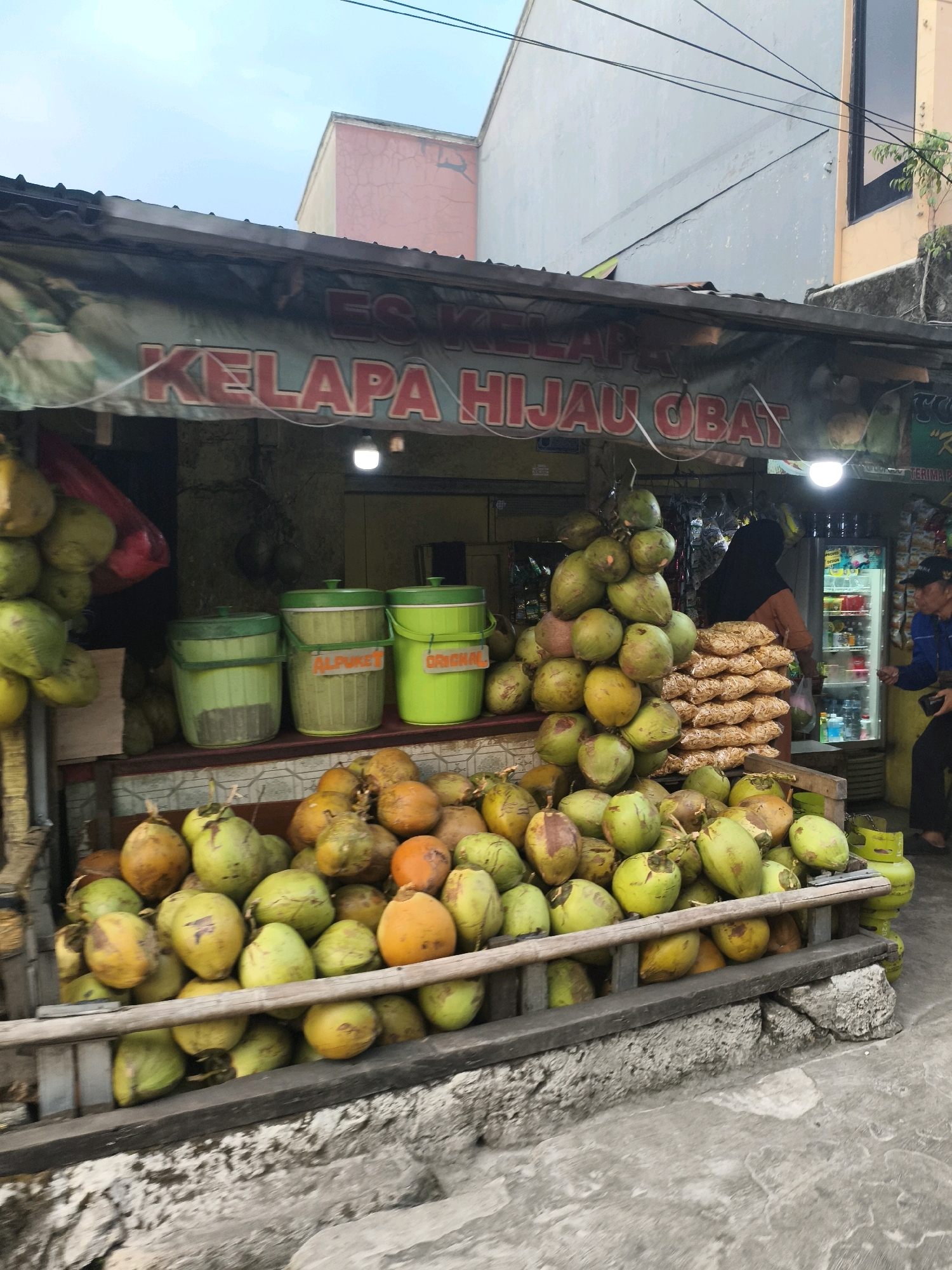 A street vendor stall in Depok, Indonesia, displays a large quantity of young green coconuts. The stall has a red and white banner overhead that reads "ES KELAPA HIJAU OBAT". Three large green plastic buckets are stacked on a shelf behind the coconuts; one has an orange label that says "ALPUKET" and another has an orange label that says "ORIGINAL". A stack of packaged snacks is visible next to the coconuts. A person wearing a black hijab and an orange top is standing behind the counter, facing the snacks. To the right, a person in a blue jacket and a black cap is looking at a refrigerated display case. The ground in front of the stall is concrete, and a stone-like structure is visible. Coconuts are piled high on the stall and on the ground.