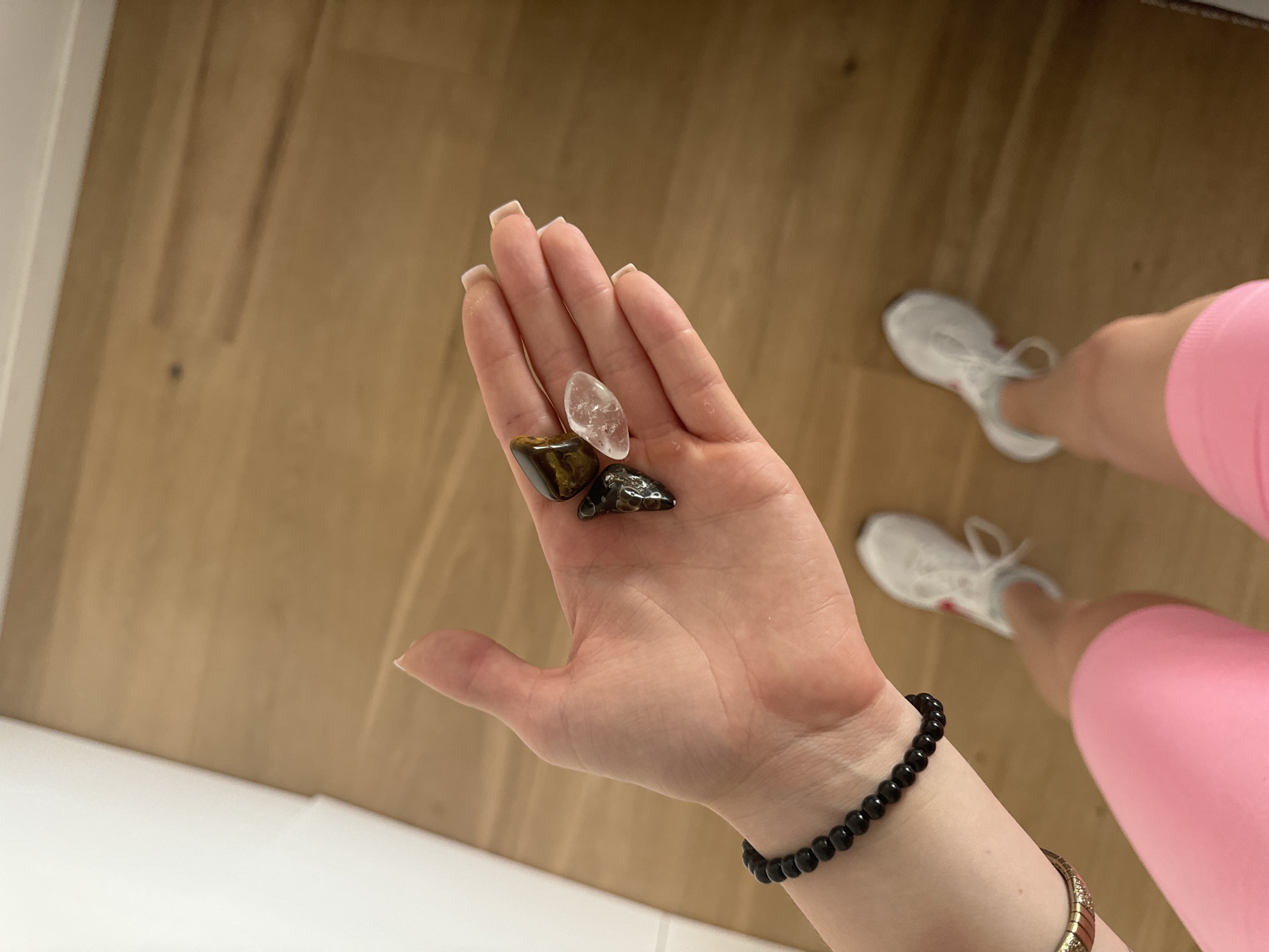A person's hand, held open with the palm facing upwards, displays three small, tumbled stones. The stones are a dark, almost black, speckled rock, a banded brown and gold stone resembling tiger's eye, and a translucent, clear quartz crystal. The hand is adorned with a black beaded bracelet on the wrist and a gold, textured bracelet partially visible below it. The background consists of a light-colored wooden floor. The person is wearing white athletic shoes and bright pink shorts. The scene appears to be indoors, possibly in a residential setting, and there are no discernible landmarks that would place it specifically in the city of Murcia, Spain.