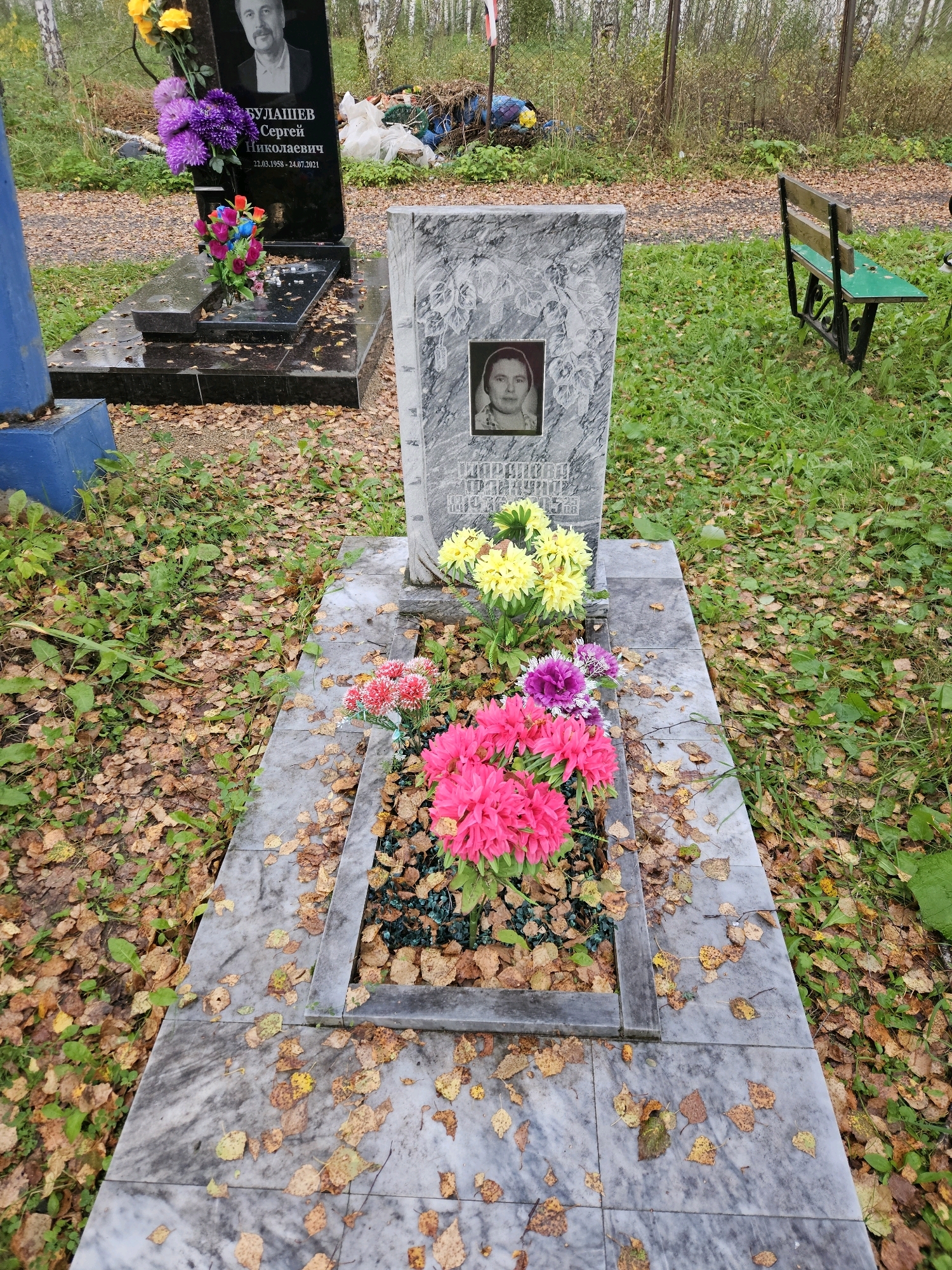 This is a photograph of a cemetery in the city of Sipavskoye, Russia. In the foreground, there is a grave marker made of marble with a framed black and white portrait of a woman. Artificial flowers in pink, red, yellow, and purple are placed in front of the marker, some in a small flower bed. Another grave marker is visible to the left, a black granite monument with a portrait of a man and Russian text inscribed. The ground around the graves is covered with fallen autumn leaves and green grass. In the background, a wooden bench with a green seat is visible, along with a fence and trees. There are also some piles of trash and debris visible in the background, suggesting the cemetery may be poorly maintained. The overall impression is one of a somber and somewhat neglected burial site.