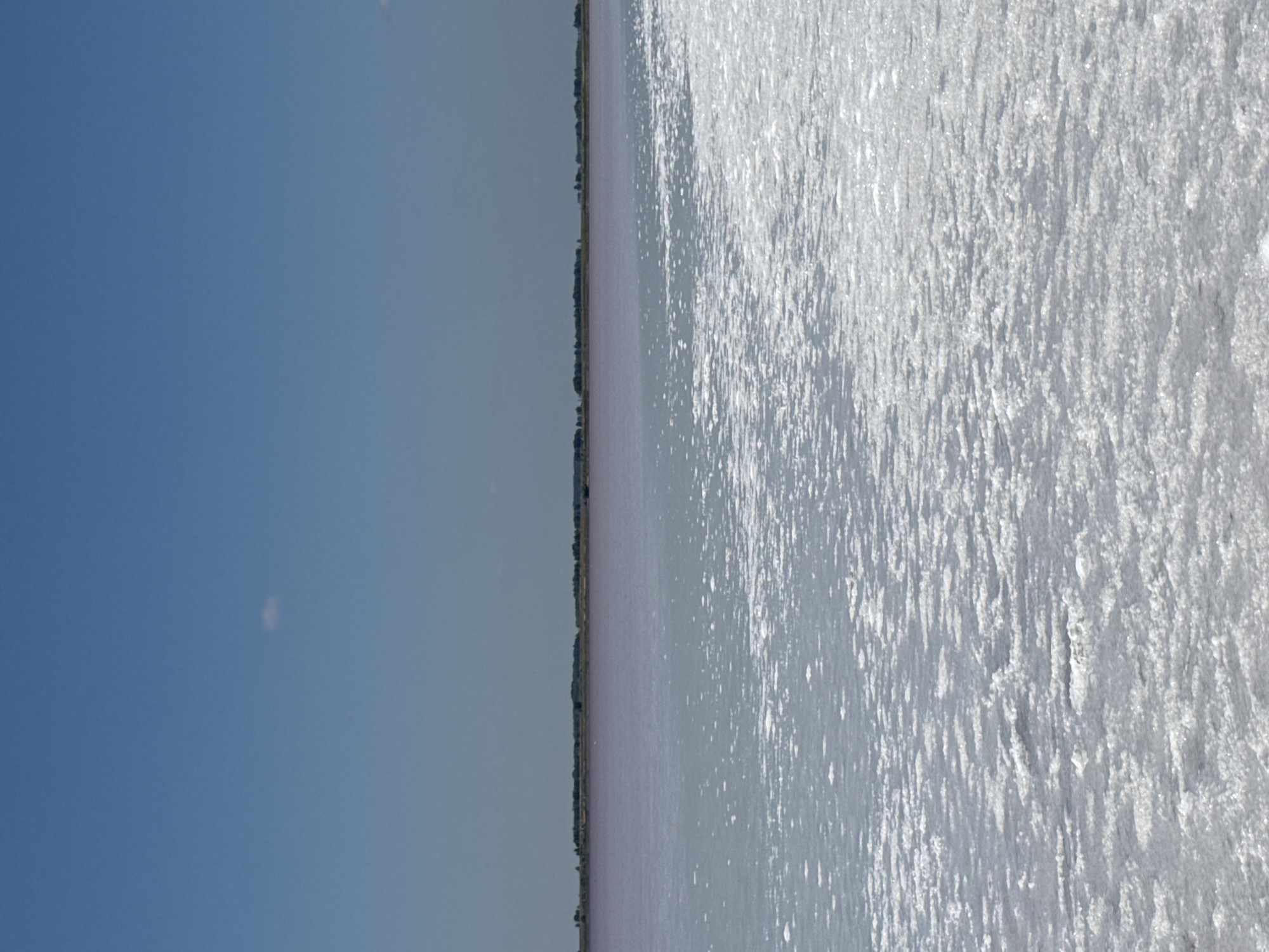 The image shows a salt lake under a clear blue sky with a single small white cloud. The foreground is dominated by a textured surface of salt crystals, reflecting light. Beyond the salt flats, there is a body of water with a pinkish hue, and a distant tree line with a few buildings visible. The scene appears to be a natural landscape, likely a salt flat or lake, in Bursol', Russia. There are no people or animals visible in the image.