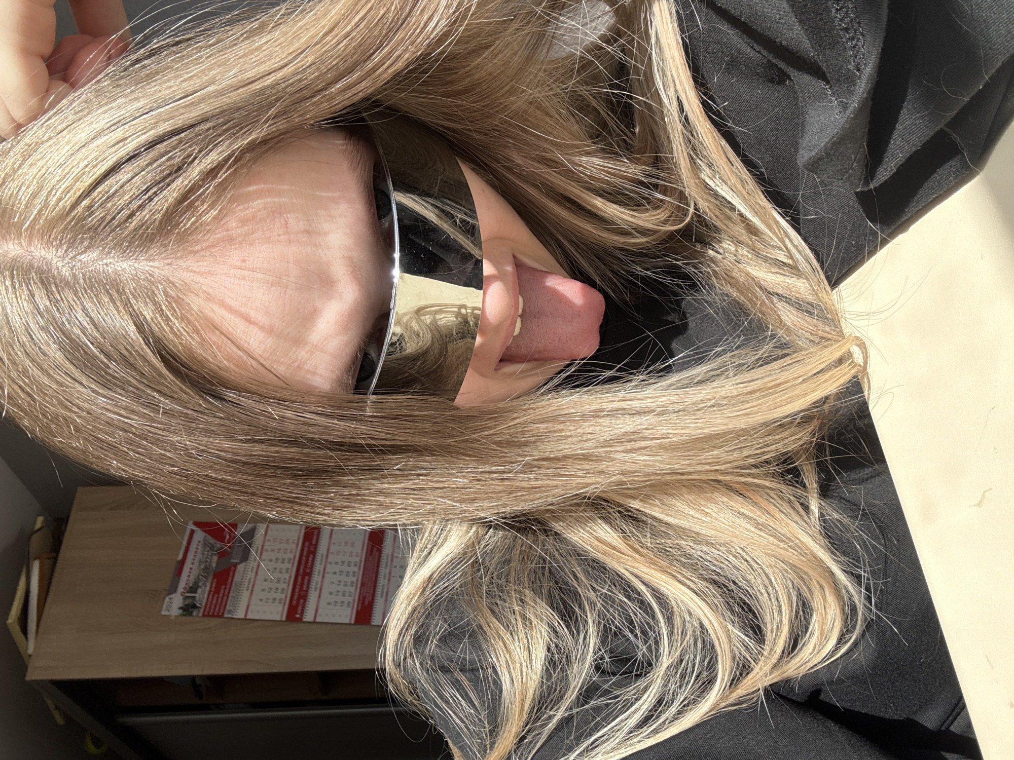 A selfie taken indoors shows a person with light-colored hair, sunglasses, and their tongue sticking out. The person is wearing a dark-colored top. A calendar and a wooden surface are visible in the background. The location is not discernible from the image.