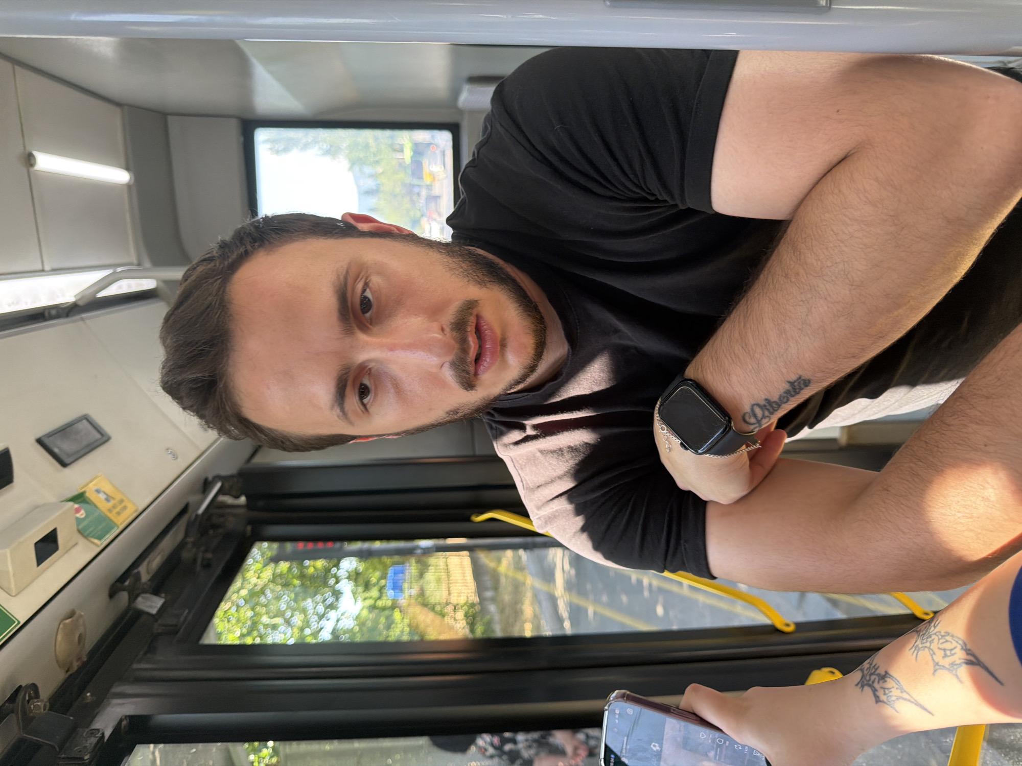 A man with a beard and short dark hair, wearing a black t-shirt, sits inside a bus in Tbilisi, Georgia. He is looking towards the camera with a neutral expression. His left arm is crossed over his chest, revealing a tattoo on his forearm that reads "Liberta." He is also wearing a smartwatch on his left wrist. To his left, another person's arm is visible, holding a smartphone. The background shows the interior of the bus, including windows with greenery visible outside, and yellow handrails. The lighting suggests it is daytime.