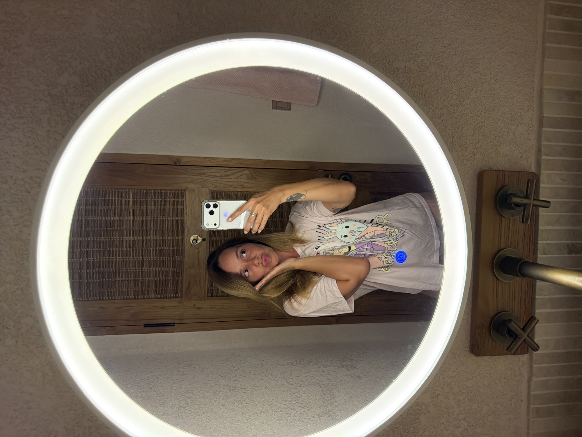 A woman takes a mirror selfie in a bathroom. She is looking at her phone, which she holds in her right hand. Her left hand is on her cheek. She is wearing a light-colored t-shirt with a cartoon graphic on it. The bathroom has a round mirror with a bright LED light around the edge. The mirror reflects a wooden door and a pink towel hanging on the wall. Below the mirror, there is a wooden shelf with two faucets. The walls of the bathroom appear to be textured. The location is a bathroom in Mengwi, Indonesia.
