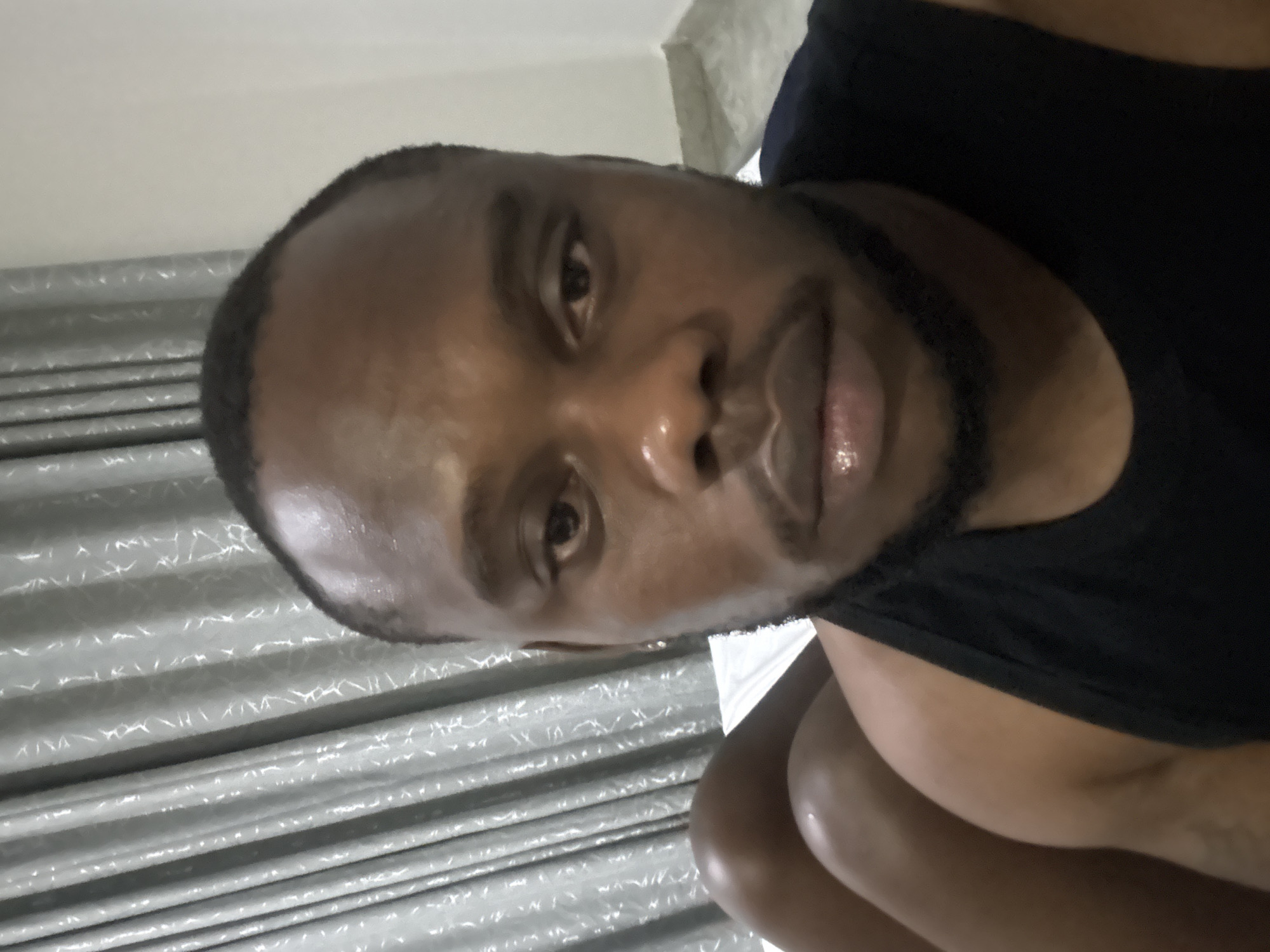 A close-up, rotated portrait image shows an adult male of African descent, likely taken indoors in Iddo Tudunwada, Nigeria. The man, with a neatly trimmed dark beard and short hair, looks directly at the camera with a neutral expression. His skin has a slight sheen, particularly on his forehead and lips. He is wearing a dark, possibly black, sleeveless top.
The background consists of a plain, light-colored wall or ceiling at the top, and a textured, light grey surface with distinct horizontal lines below it, which could be blinds, corrugated metal, or patterned curtains. The lighting appears to be indoor or artificial. The image is rotated 90 degrees counter-clockwise, presenting the man's head tilted to his right. There is no visible text.