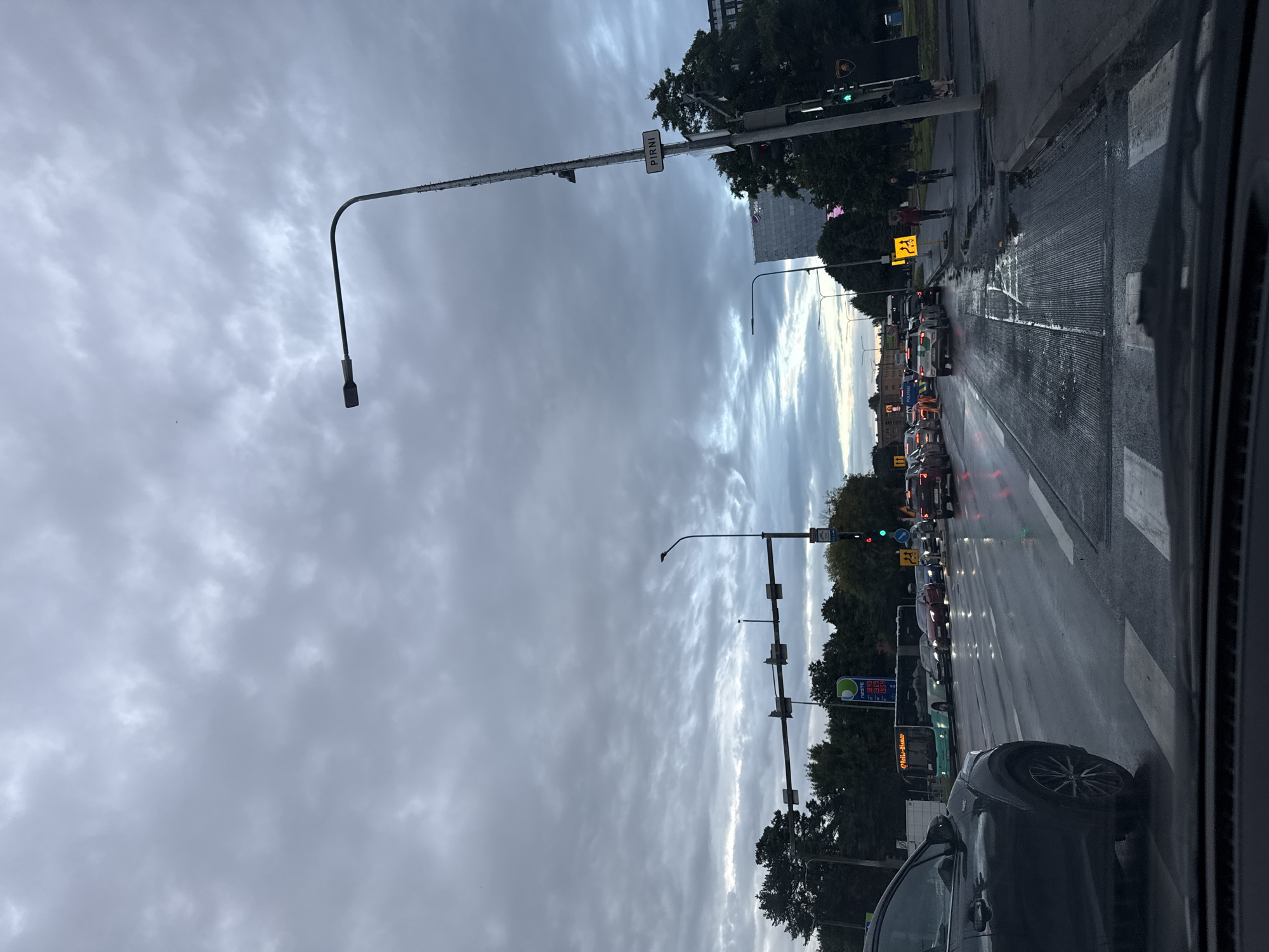 The image shows a cloudy sky over a street in Tallinn, Estonia. In the foreground, a car is stopped at a pedestrian crossing. The wet road surface reflects the streetlights and the headlights of other vehicles. Traffic is backed up, with several cars and a bus visible. A traffic light is visible, showing a green signal. A sign with the word "PIRNI" is mounted on a lamppost. In the background, trees line the street, and a tall building can be seen. A gas station with prices displayed is also visible. Several people are walking and cycling on the sidewalk. The overall impression is of a busy urban street scene on a cloudy day.