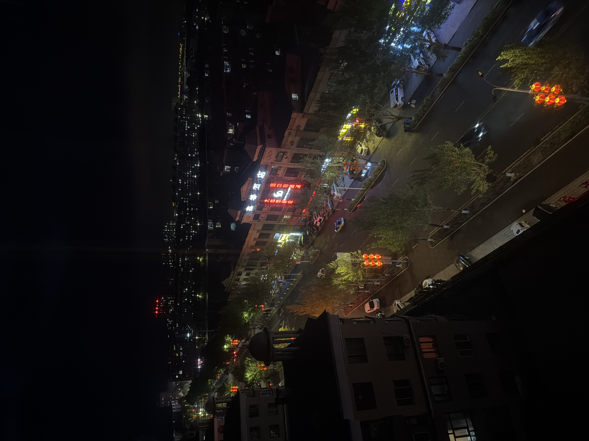 This is a nighttime aerial view of a bustling city street in Tongjiang, China, taken from an elevated perspective. The frame is dominated by a dark night sky on the left, contrasting with the illuminated urban environment on the right.
In the midground, a multi-lane road stretches diagonally across the image. It is lined with numerous trees, some silhouetted against the night sky, while others are brightly lit by streetlights. Several vehicles are visible on the road, some appearing as blurred streaks, indicating movement. Distinctive traffic lights, designed as stacks of red lanterns, are prominently displayed at various points along the street, casting a warm red glow.
A prominent, multi-story building stands alongside the street, featuring a large, brightly illuminated sign. The Chinese characters "古贺" (Gǔhè) are at the top, followed by "汤泉宾馆" (Tāngquán Bīnguǎn), which translates to "Hot Spring Hotel." Below this, the English text "K-BOWLING" and "GO" is also visible. Many windows in this and other surrounding buildings are lit, suggesting ongoing activity.
Further into the background and along the left edge, more buildings extend into the distance, their scattered lights twinkling. The foreground shows the dark roof and upper sections of a building from which the photo appears to have been taken. The scene captures the typical vibrant life of a city at night, with artificial lights defining the urban landscape under a clear sky. No specific individuals are clearly visible.