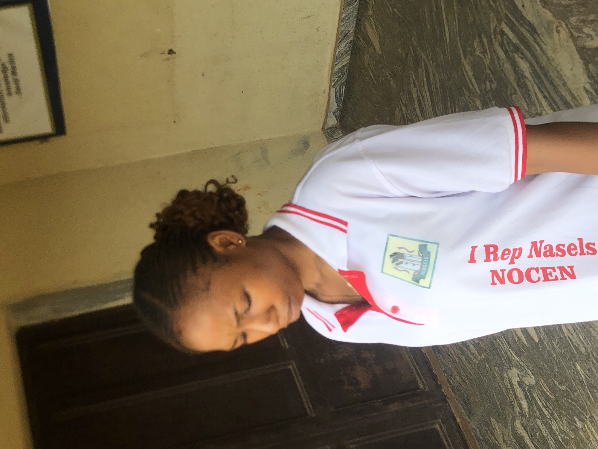 A young woman is depicted in a medium shot, oriented vertically in the image but appearing horizontally due to the original capture angle. She is positioned slightly off-center to the left, with her head and upper torso visible. She wears a white short-sleeved polo shirt with red trim on the collar, placket, and sleeve cuffs. On the right side of her chest, the shirt features a logo and text. The logo is a green, somewhat shield-shaped emblem with "NASELS" written within it, along with what appears to be a building or symbolic graphic. Adjacent to the logo, the red text "I Rep Nasels NOCEN" is printed on the shirt.
The woman has dark, curly hair styled in an updo. Her eyes are closed, and her facial expression appears serene or relaxed. A small, gold-colored stud earring is visible in her right ear.
The background consists of a light-colored, possibly beige or cream, wall on the left side, where a framed, blurry object with unreadable text hangs. To the right and below her, there's a dark, wooden-looking door or surface. The bottom right of the image shows a textured, patterned floor or wall, possibly marble or granite, with shades of brown and grey. The lighting appears to be natural, suggesting daytime. The scene is set in Nsugbe, Nigeria.