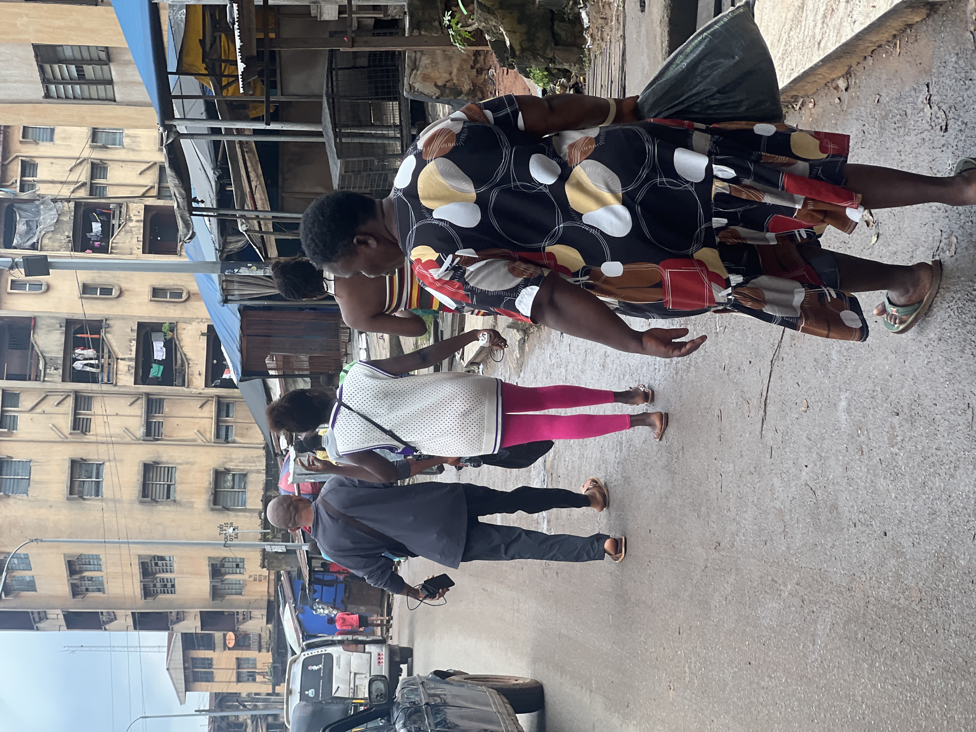 The image depicts a street scene in Nkpor, Nigeria, likely a market area given the presence of makeshift stalls and densely packed buildings. A group of people are walking away from the camera on a paved, wet road. In the foreground, a woman wearing a patterned dress and carrying a black plastic bag walks ahead of another woman wearing a white sleeveless top and bright pink leggings. Behind them, a man in a dark grey long-sleeved shirt and black trousers walks with a phone and wallet in his hand. A white van is visible to the left of the frame, and other people and market stalls are present in the background, under overcast skies. The buildings are multi-story and appear aged, with some structures showing signs of disrepair.