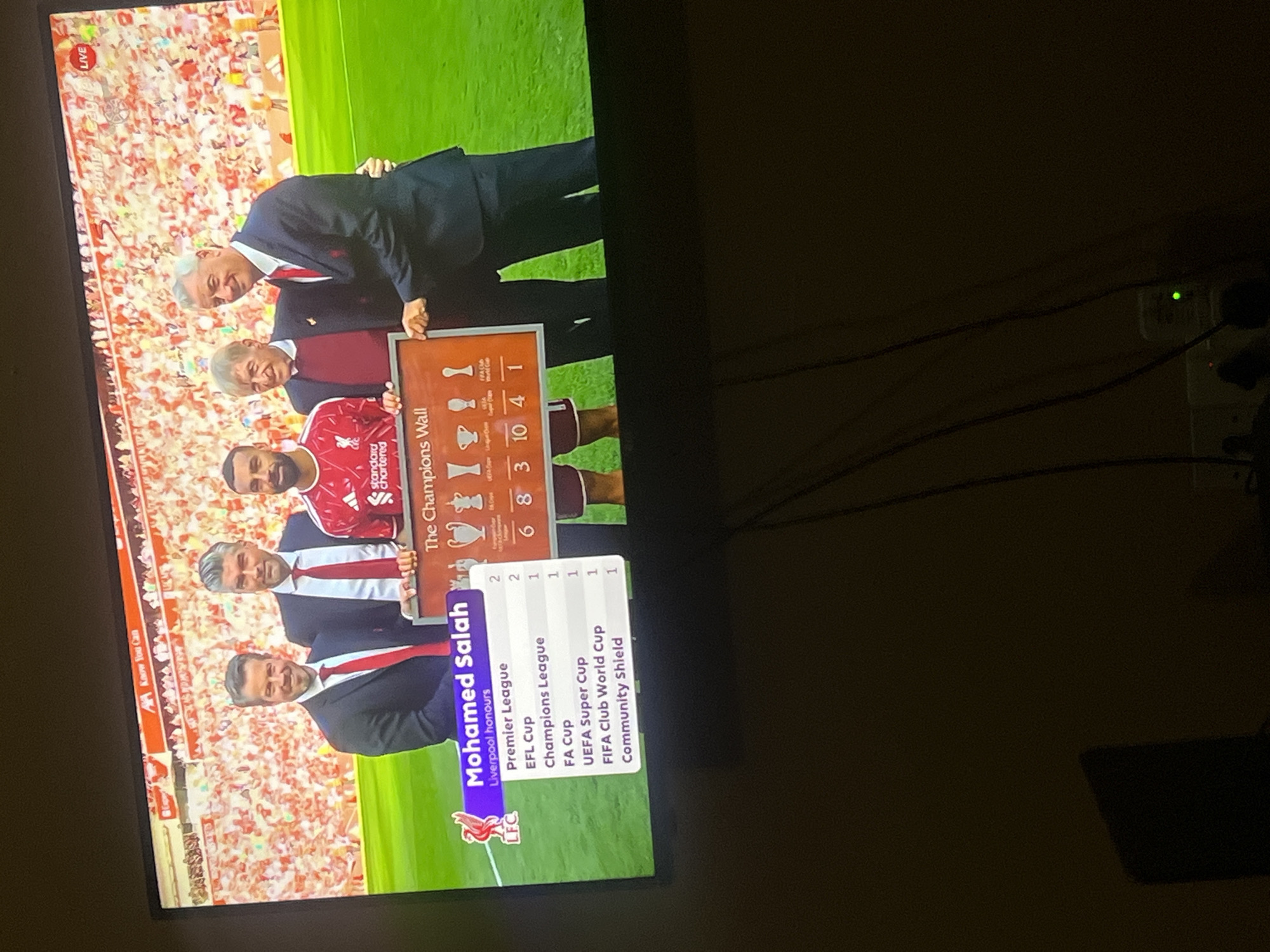 The image shows a television screen displaying a graphic about Mohamed Salah's achievements with Liverpool FC. The graphic lists his honors, including Premier League titles, FA Cups, Champions League titles, and others. Salah is pictured in his Liverpool kit. People in suits are also shown holding up a Champions Wall with trophies listed. The background is a soccer stadium with fans. The room is dimly lit and the image is taken at an angle. There is a white power outlet with several cords coming from it visible in the right. No location in Oshogbo, Nigeria is evident in the image.