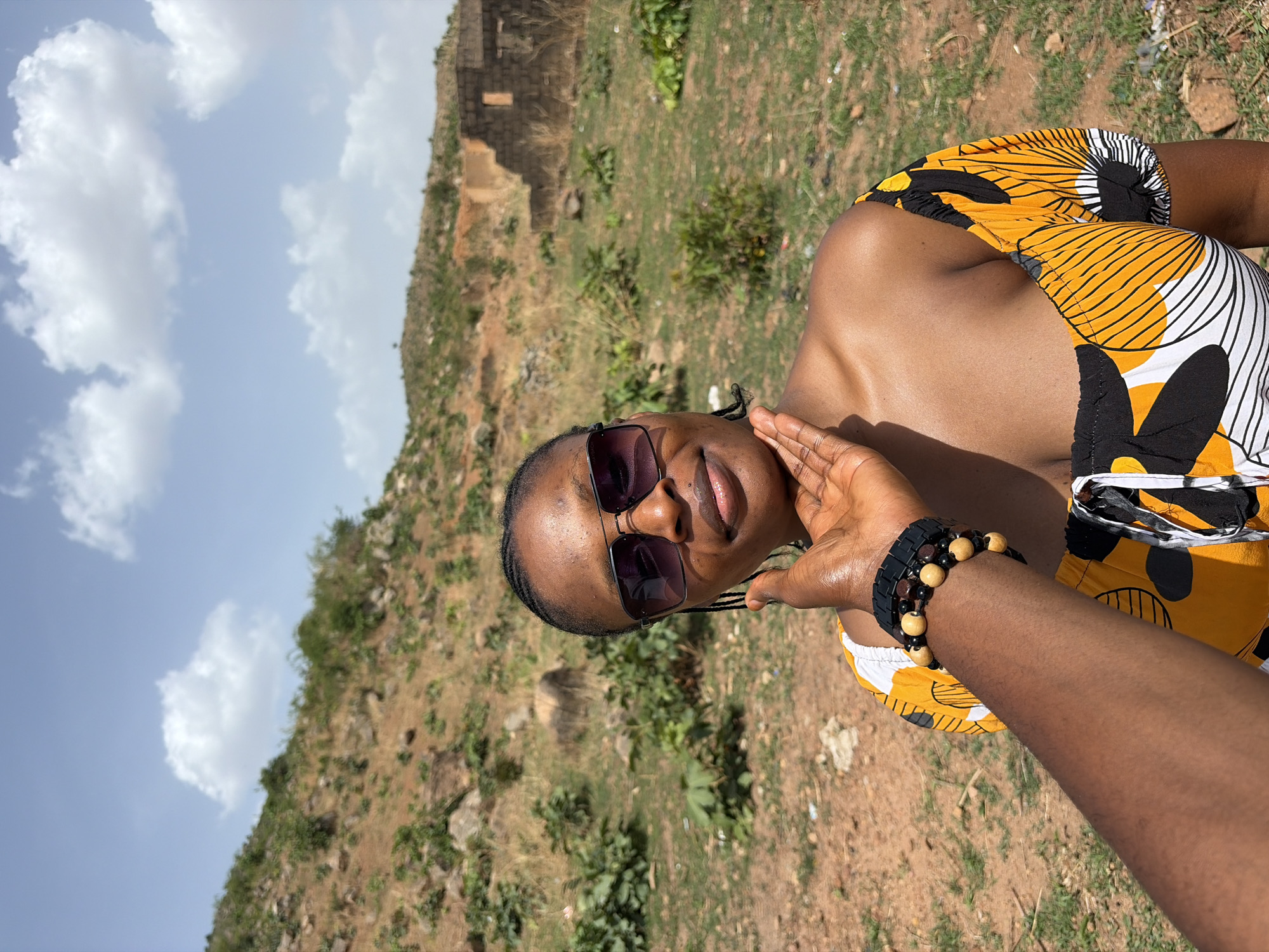 The image captures a smiling woman outdoors on a bright, sunny day, set against a rocky, vegetated hillside under a blue sky with scattered clouds. The woman is positioned in the foreground, seen from the chest up, wearing dark sunglasses and a vibrant yellow, black, and white patterned top, characteristic of African print. Her right hand is gently raised to her face, partially obscuring her cheek, and her wrist is adorned with a stack of bracelets featuring dark and light wooden beads. Her skin appears moisturized or glistening in the sunlight.
In the background, a dry, rugged landscape unfolds, characterized by reddish-brown earth, sparse green shrubs, and rocks. A weathered stone structure, possibly ruins, can be seen on the upper part of the hill. The bright light suggests it is daytime, likely mid-day or early afternoon. A segment of an arm, presumably the photographer's, is visible in the lower right corner, indicating a close-up shot or a selfie. The overall atmosphere conveys relaxation and enjoyment in an outdoor, natural setting, consistent with the location context of Jos, Nigeria. No visible text is present in the image.