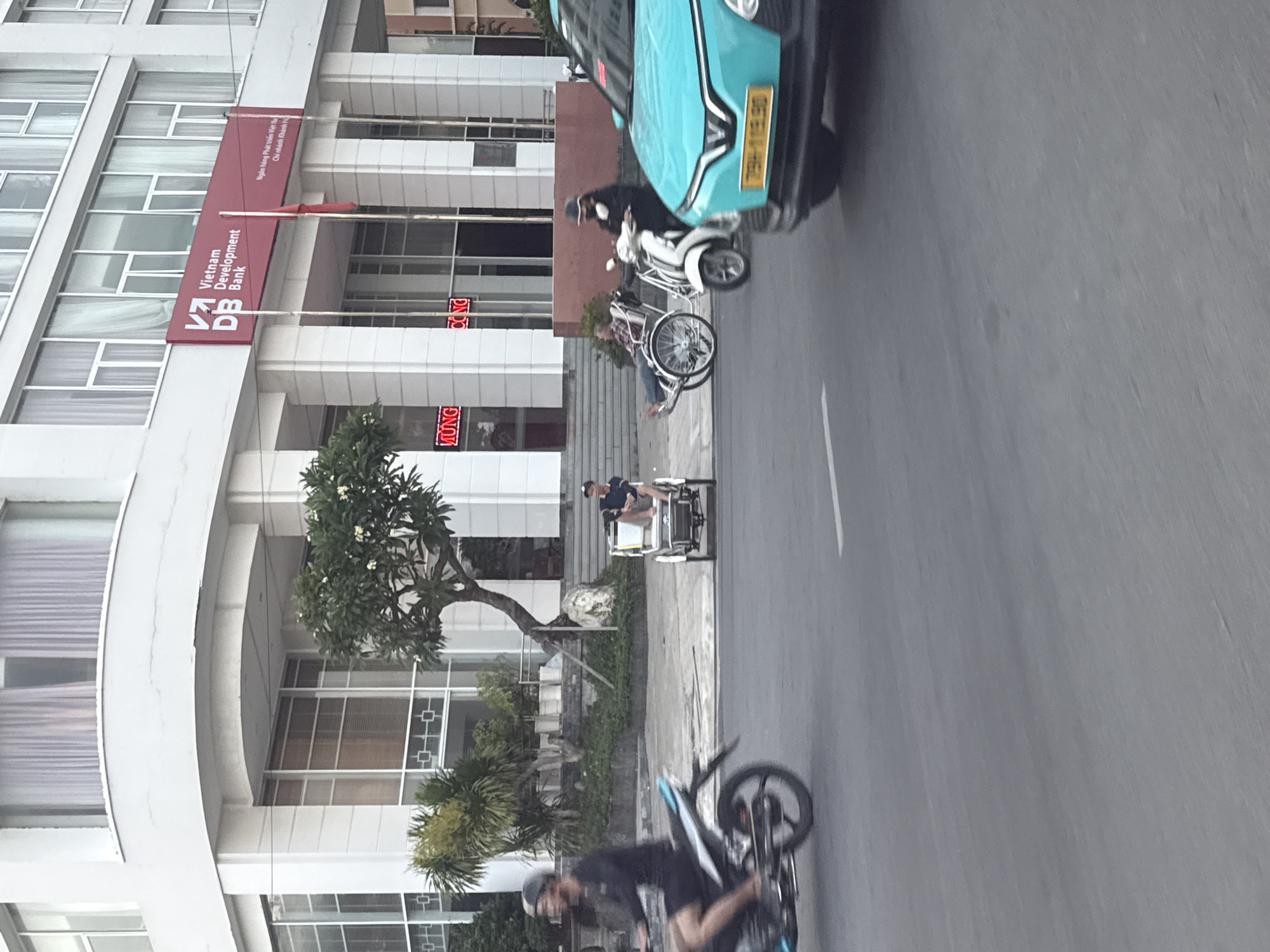 A busy street scene unfolds in front of a modern building with a red sign reading "Vietnam Development Bank." A man on a motorcycle is captured in motion on the left side of the frame, while a person in a wheelchair is stationary in the middle ground. A light blue car is prominent on the right, partially obscuring the street. The overall environment suggests an urban setting in Vietnam, possibly Bac Nha Trang Ward, based on the presence of Vietnamese signage and vehicles. The image appears to be taken during daylight hours, with a slight overcast sky.
