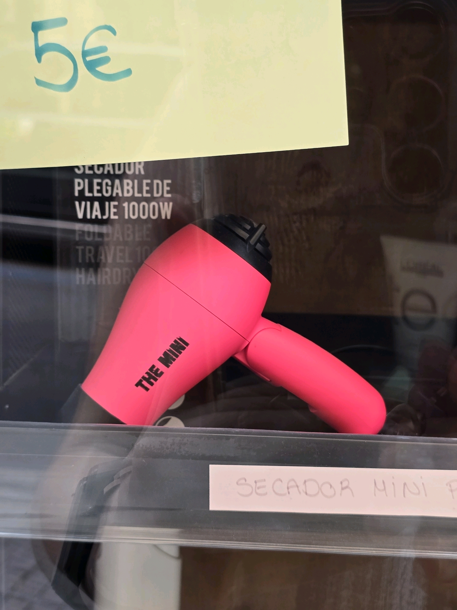 A bright pink compact hairdryer, labeled "THE MINI", is displayed behind a clear plastic barrier, possibly in a shop window or vending machine, in Jaén, Spain. The hairdryer rests on a dark surface, and is partially illuminated by a pink light source. Above the hairdryer, a yellow sign displays "5€" in large, blue handwritten text. Below the hairdryer, another white sign reads "SECADOR MINI P". The background is a dark, reflective surface with hints of other products, including a blurred product with the "L'ORÉAL" logo visible.