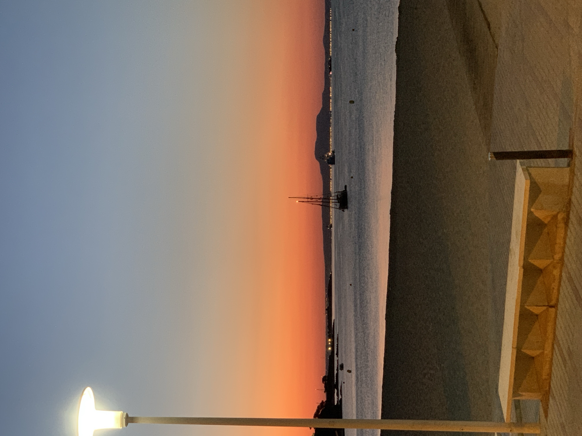 This image depicts a serene sunset scene on the beach in Palmanova, Spain. In the foreground, a long, concrete bench sits on a paved walkway bordering a sandy beach. A modern lamppost with a glowing light stands to the left, casting a warm illumination. The tranquil sea stretches out to the horizon, reflecting the vibrant hues of the sunset, which paints the sky in shades of orange and blue. Several boats, including a sailboat with tall masts and a larger ship, are visible on the water. In the distance, a silhouetted landmass, possibly an island or coastline, is dimly visible, with scattered lights suggesting human habitation. The overall atmosphere is peaceful and picturesque, capturing the beauty of the coastal landscape at dusk.