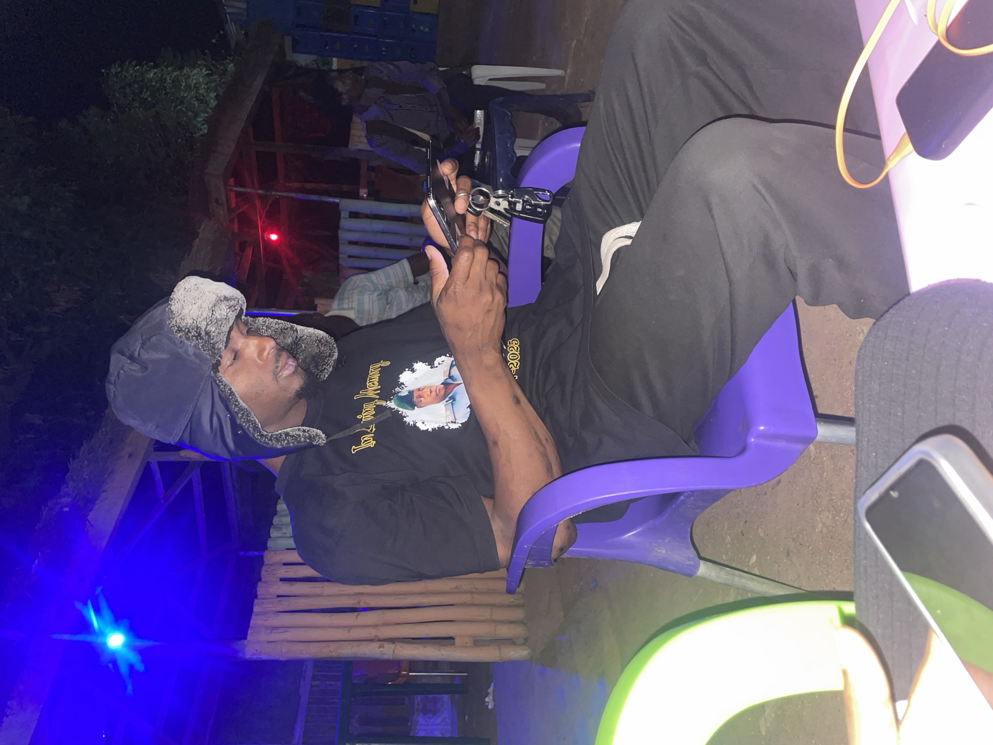 A man wearing a dark-colored trapper hat and a black t-shirt with a graphic print is seated on a purple plastic chair. He is holding a set of keys and a nail clipper. His legs are covered in dark pants. To his right, there's a light green plastic chair and a mobile phone. The area is illuminated by a blue light source in the bottom left corner. Behind him, there are other people seated on white plastic chairs. A red light is visible in the background. The setting appears to be outdoors, and could be in Jos, Nigeria.