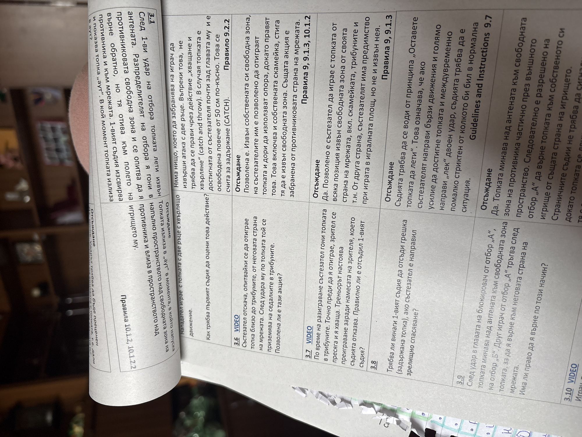 The image shows a close-up of a document, likely a rulebook or guidelines, pertaining to volleyball. The text is in Bulgarian, and it covers different scenarios and interpretations of rules, with titles like "Отсъждане" (Judging) and "VIDEO," indicating potential video examples. Topics include hitting the ball, player actions around the net and sidelines, interference, and the application of rules in different situations. There are also references to specific rule numbers (e.g., Правило 9.2.2, Правила 9, 9.1.3, 10.1.2, Правила 10.1.2, 10.1.2.2) and guidelines from official documents. The document is lying on a slightly reflective surface, possibly a table, with a small portion of what appears to be a hand or another object visible at the bottom of the image, near some colorful dots.