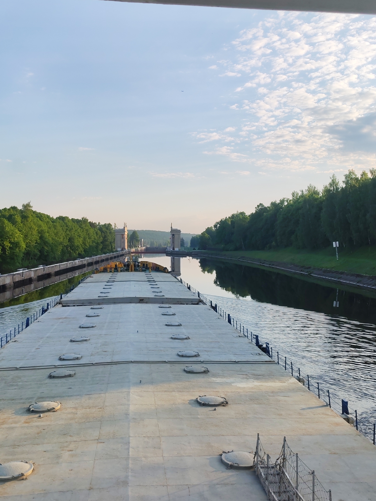 The image depicts a large, flat-decked barge traversing a canal in what appears to be a natural setting. The location is likely within or near the city of 3-Y Uchastok, Russia.
The barge occupies the foreground and extends into the midground. Its deck is composed of a series of rectangular panels, punctuated by numerous circular access hatches. A railing runs along the edge of the barge, separating the deck from the canal.
The canal, flanked by dense green trees on both sides, stretches toward a structure in the distance that could be a lock or other canal control facility. The sky above is partially cloudy, with a blue expanse visible between the clouds. The water in the canal is relatively calm, with reflections of the sky and surrounding trees.