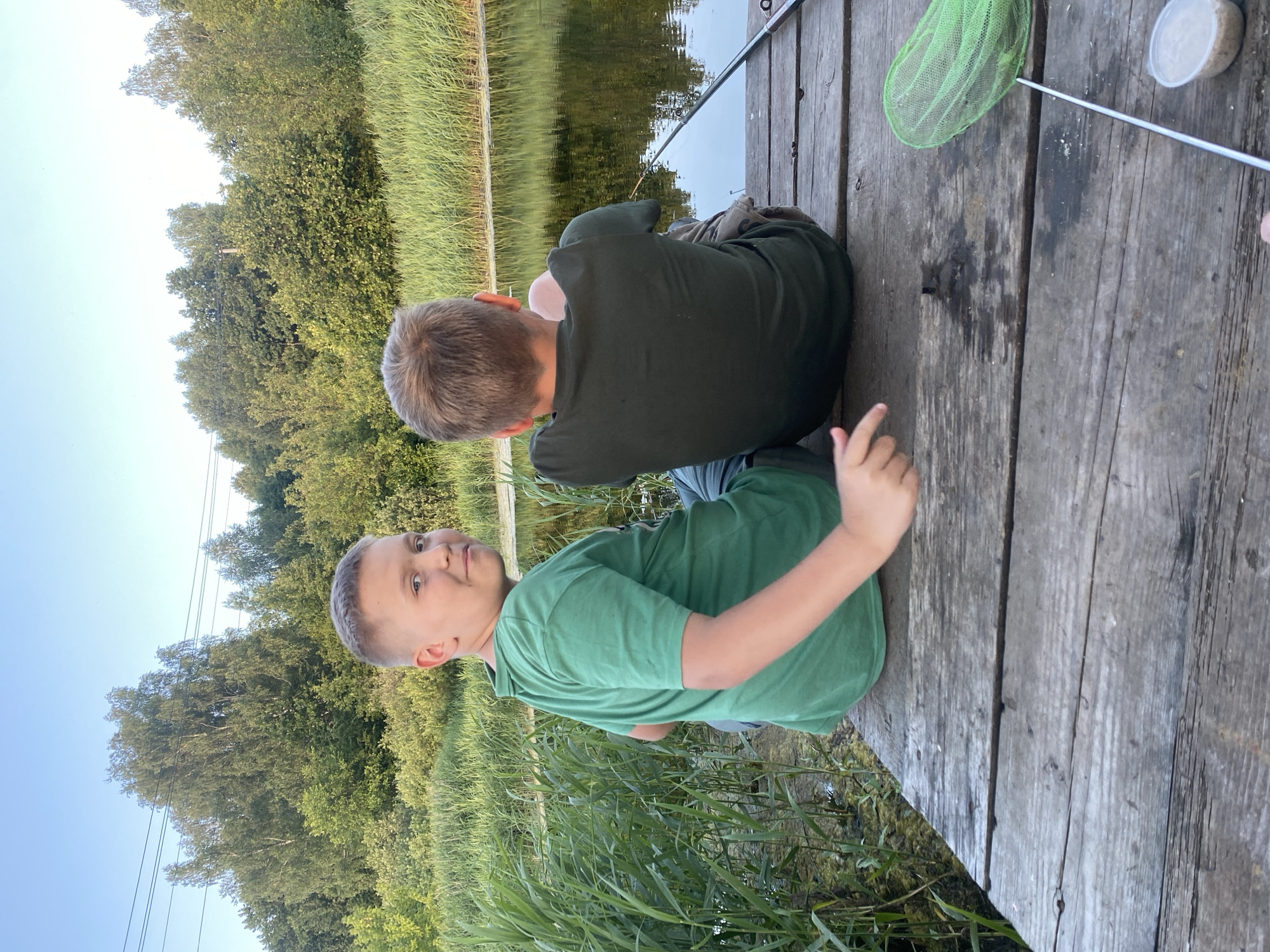 Two young boys are sitting on a wooden dock facing away from the camera, towards a body of water. The boy on the left, wearing a green t-shirt, is looking back at the camera and smiling. He is positioned on the left side of the dock. The boy on the right, wearing a dark green t-shirt, is looking towards the water, with his arms resting behind him. A fishing rod lies on the dock, extending into the water. A green fishing net and a small white container are also visible on the dock. The background consists of lush green trees and tall reeds bordering the water. The sky is a clear, pale blue. The scene appears to be outdoors in a natural setting, possibly in the village of Afanasyevo, Russia.