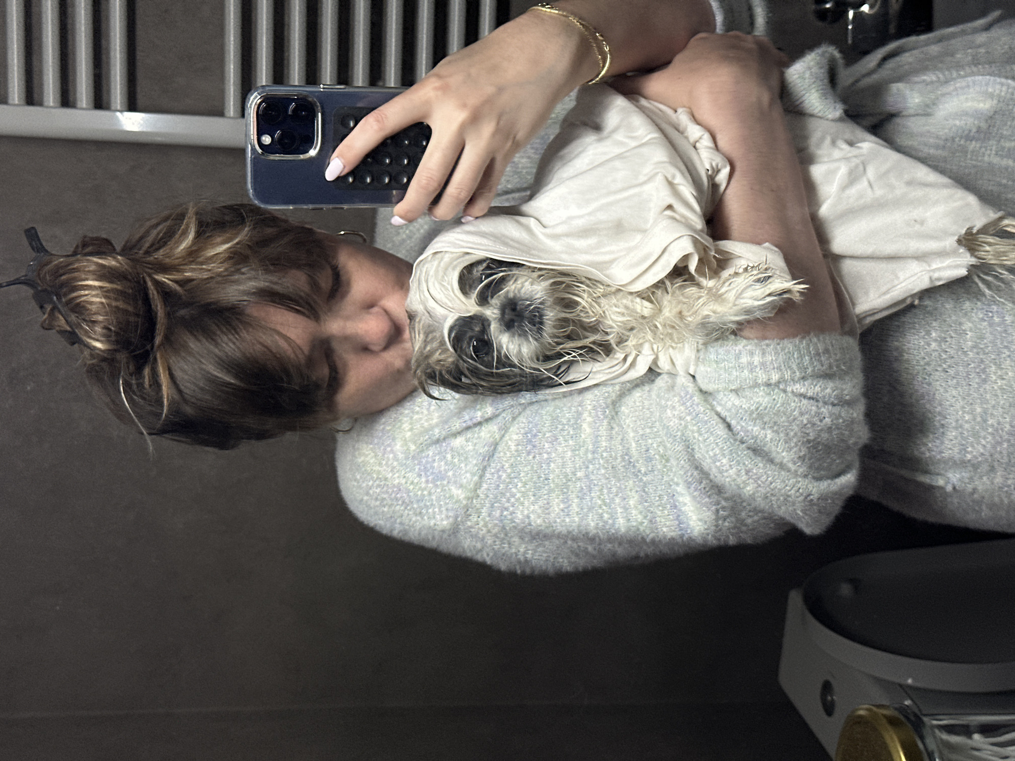 A person with light brown hair pulled back in a messy bun, wearing a light-colored sweater, is holding a small dog wrapped in a white towel. They are taking a selfie in front of a mirrored wall with vertical metal bars. The person's face is close to the dog's. The phone, with a blue case and multiple camera lenses, is in their hand. A gold bracelet is visible on their wrist. The lower right corner of the image shows a scale with a gold-colored dial. The location is likely a bathroom setting. No indication of Tbilisi, Georgia is apparent from the image.