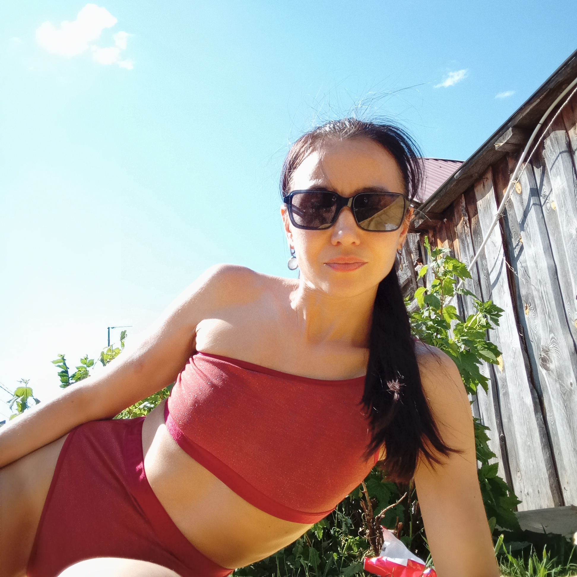 A woman with dark hair and sunglasses is lying on the grass in front of a wooden fence and greenery. She is wearing a maroon-colored two-piece outfit. In the background, there is a blue sky with a few clouds, and part of a wooden structure with a red roof is visible behind the fence. The location is likely in a rural or suburban area of Буренка, Russia, given the presence of the wooden fence, greenery, and a partially visible wooden building.