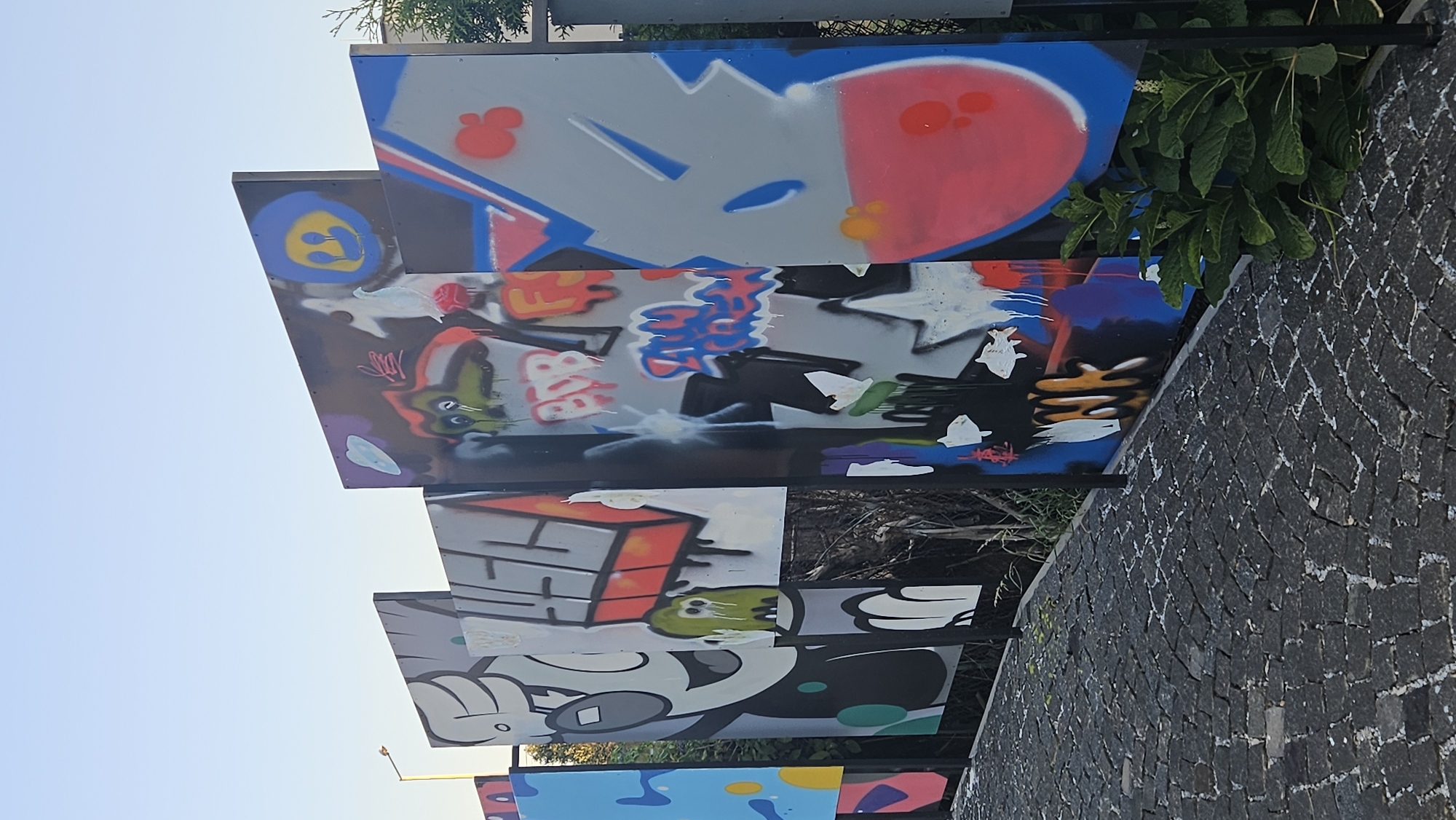The image displays a row of large, rectangular panels covered in vibrant graffiti art, set against a clear sky. The panels are arranged in a staggered formation, creating a dynamic visual. The graffiti features a variety of styles, including cartoon characters, abstract shapes, and stylized lettering, rendered in bold colors like red, blue, yellow, and pink. Some panels depict playful figures, such as a smiling cartoon character with glasses and a blue smiley face with tears. Other sections are covered in abstract designs and graffiti tags. The panels are mounted on black metal frames, and a black metal fence runs along the bottom of the scene. In the foreground, a textured cobblestone pathway stretches towards the right of the image. Lush green foliage is visible at the base of the panels on the right side, and a faint silhouette of a street light can be seen on the left. The overall impression is a public art installation.
The graffiti panels are located at the street art installation at the Sándor Petőfi Slovak Theatre in Prešov, Slovakia. This outdoor exhibition showcases contemporary street art and is a notable feature of the theatre's public space.