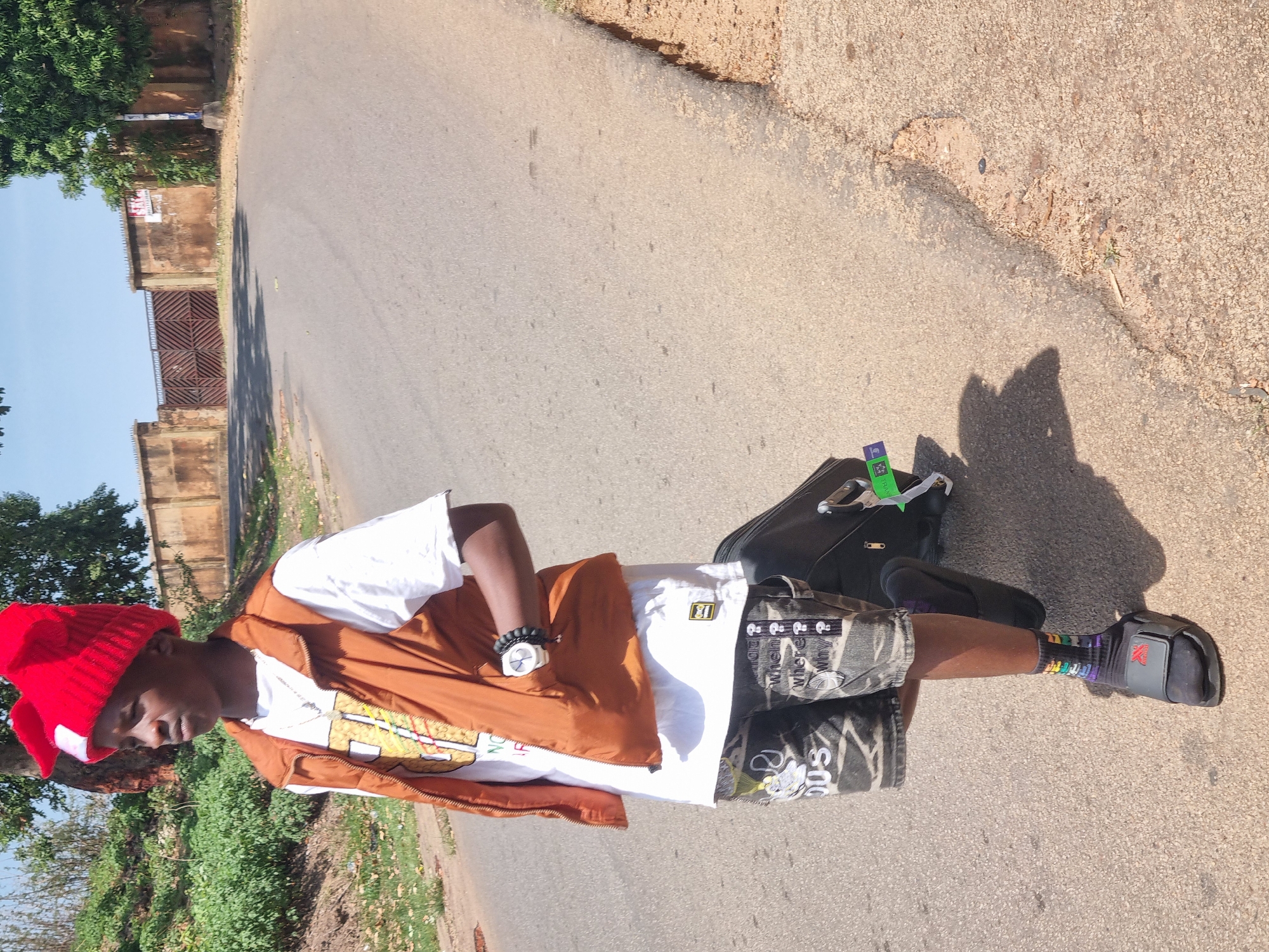 A young man stands on an asphalt road, likely in Jos, Nigeria, based on the visual cues of the environment and clothing. He wears a red knit cap, a white t-shirt with a colorful design, a brown vest, patterned shorts, and black sandals with socks that have a rainbow color pattern on his left leg. A black bag rests beside him. The road extends into the background, flanked by green foliage, trees, and building walls. The overall scene is bathed in sunlight.