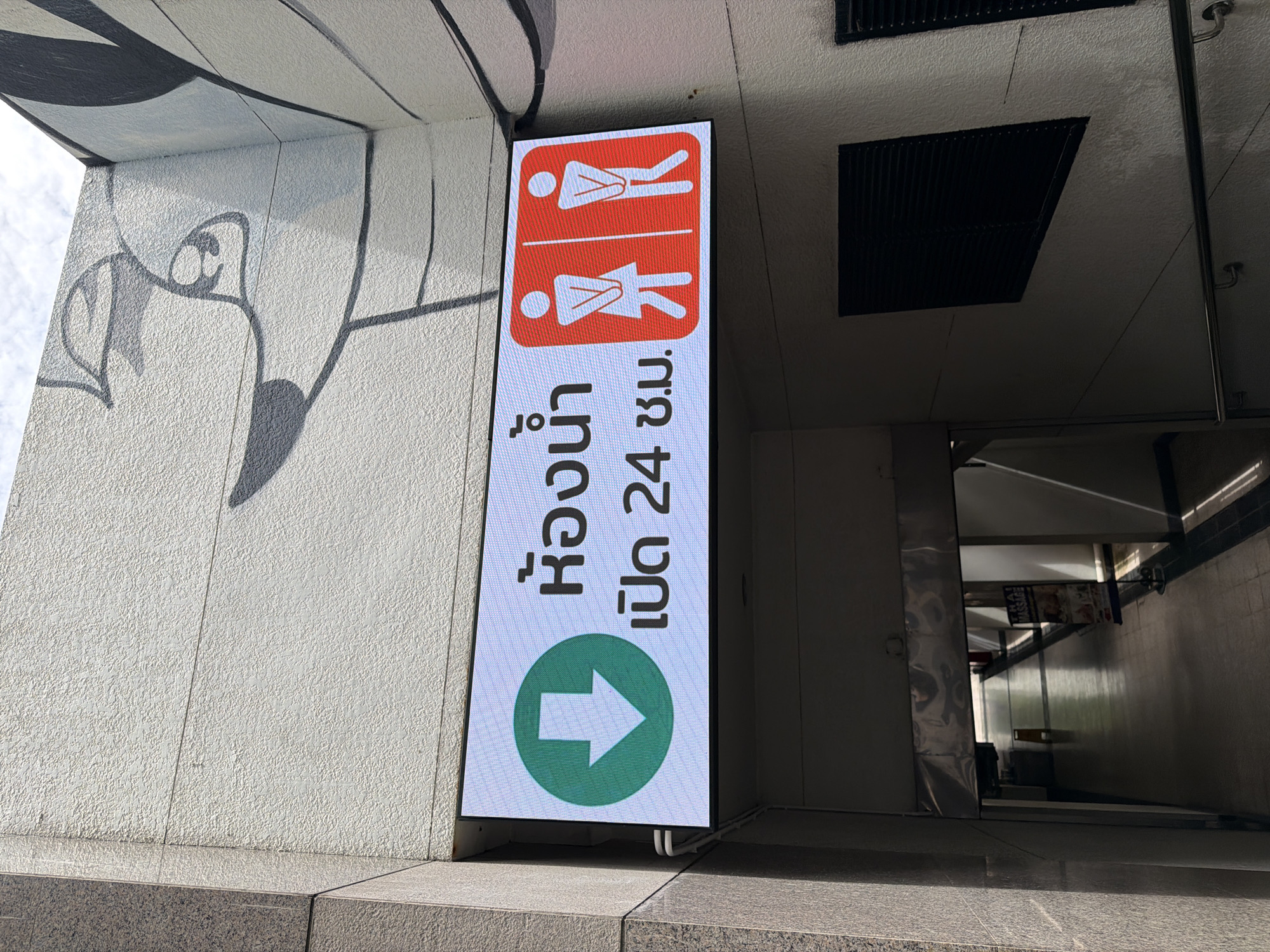 The image displays a digital sign above an entrance to an underground walkway in Bangkok, Thailand. The sign is illuminated and shows Thai text indicating "Restrooms" and the operating hours "Open 24 hours," accompanied by icons for male and female restrooms. A green downward arrow symbol is also present on the left side of the sign, suggesting the direction to the restrooms. The walkway entrance is visible below the sign, leading into a dimly lit corridor with a tiled floor. Sunlight casts long shadows down the corridor, and a sign for "Thai Massage" is partially visible on the right side. The exterior of the building features a textured white wall with a stylized mural of a face drawn in black ink.