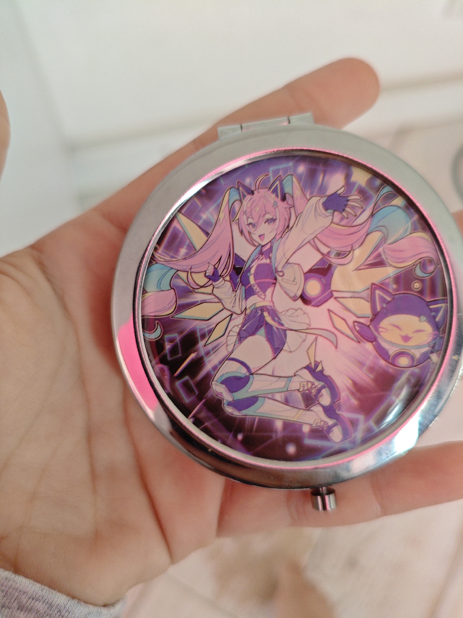A hand holds a round, silver compact mirror. The mirror's surface displays a vibrant anime-style illustration of a pink-haired female character with cat ears, wearing a futuristic purple and white outfit. She is depicted in a dynamic pose with energy lines radiating from her. To her right, a small, purple, cat-like robot with a smiley face is visible. The background of the illustration features geometric patterns and a neon aesthetic in shades of purple, blue, and yellow. The hand holding the mirror is light-skinned and appears to be wearing a grey, ribbed fabric cuff on the lower arm. The background is out of focus, showing a plain white surface with subtle shadows. There are no indications of the city of Трудовское сельское поселение, Ukraine, in this image.