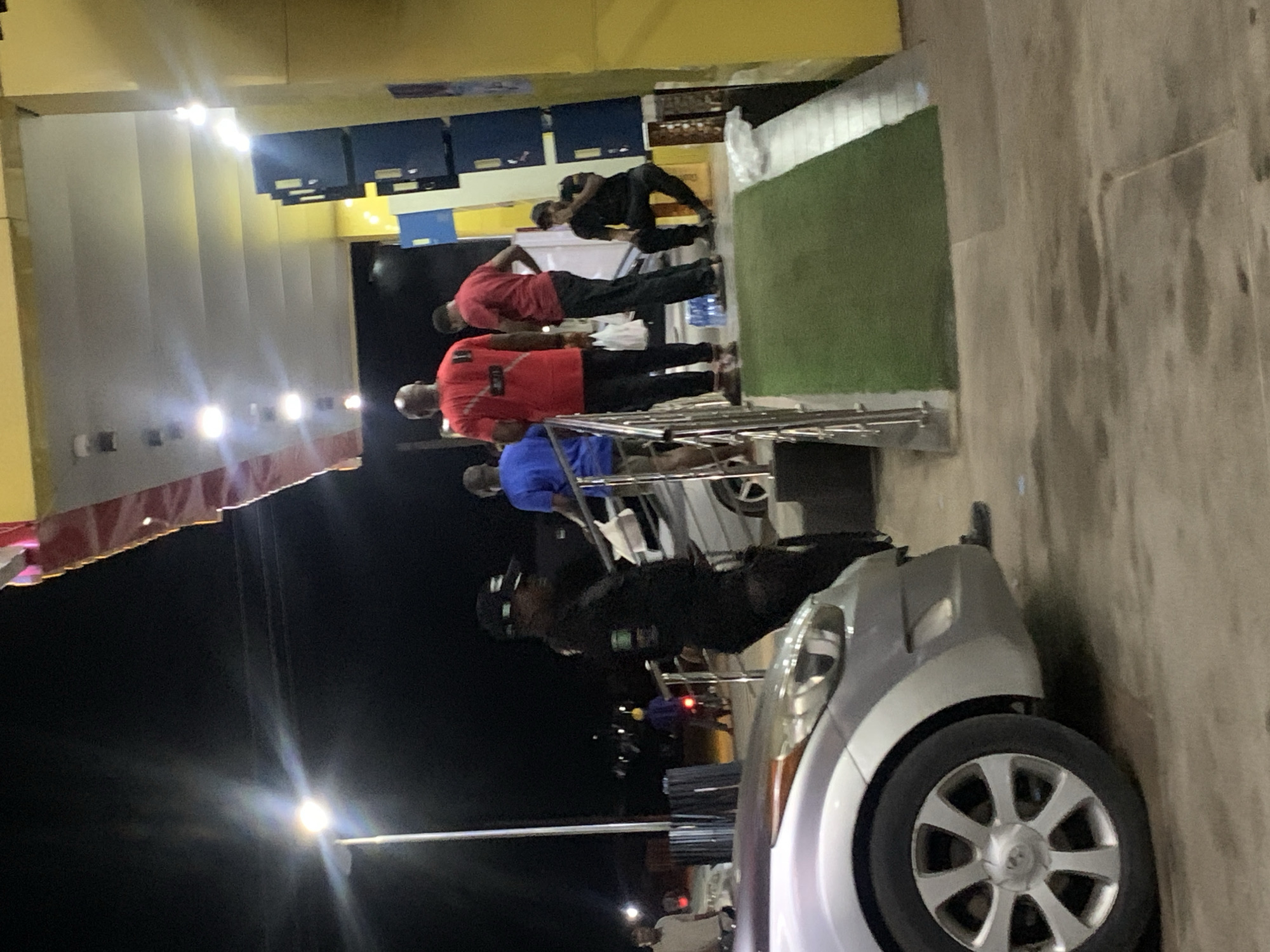 The image depicts a nighttime scene outside a building. A group of individuals are standing outside, near a car. The location is not identifiable as Abuja, Nigeria, from the visual information provided.
**People:** Several men are present, some wearing red shirts, one wearing a blue shirt, and others in dark clothing.
**Subjects:** A silver car is parked in the foreground.
**Objects:** The building in the background has a display of dark-colored boxes. There are also stairs with a metal railing and a green mat on the landing. A light pole illuminates the area.
**Actions:** The individuals are standing and talking.
**Interactions:** There appear to be casual interactions between the individuals.
**Environment:** It is nighttime. The area is illuminated by streetlights and building lights. The ground appears to be paved.