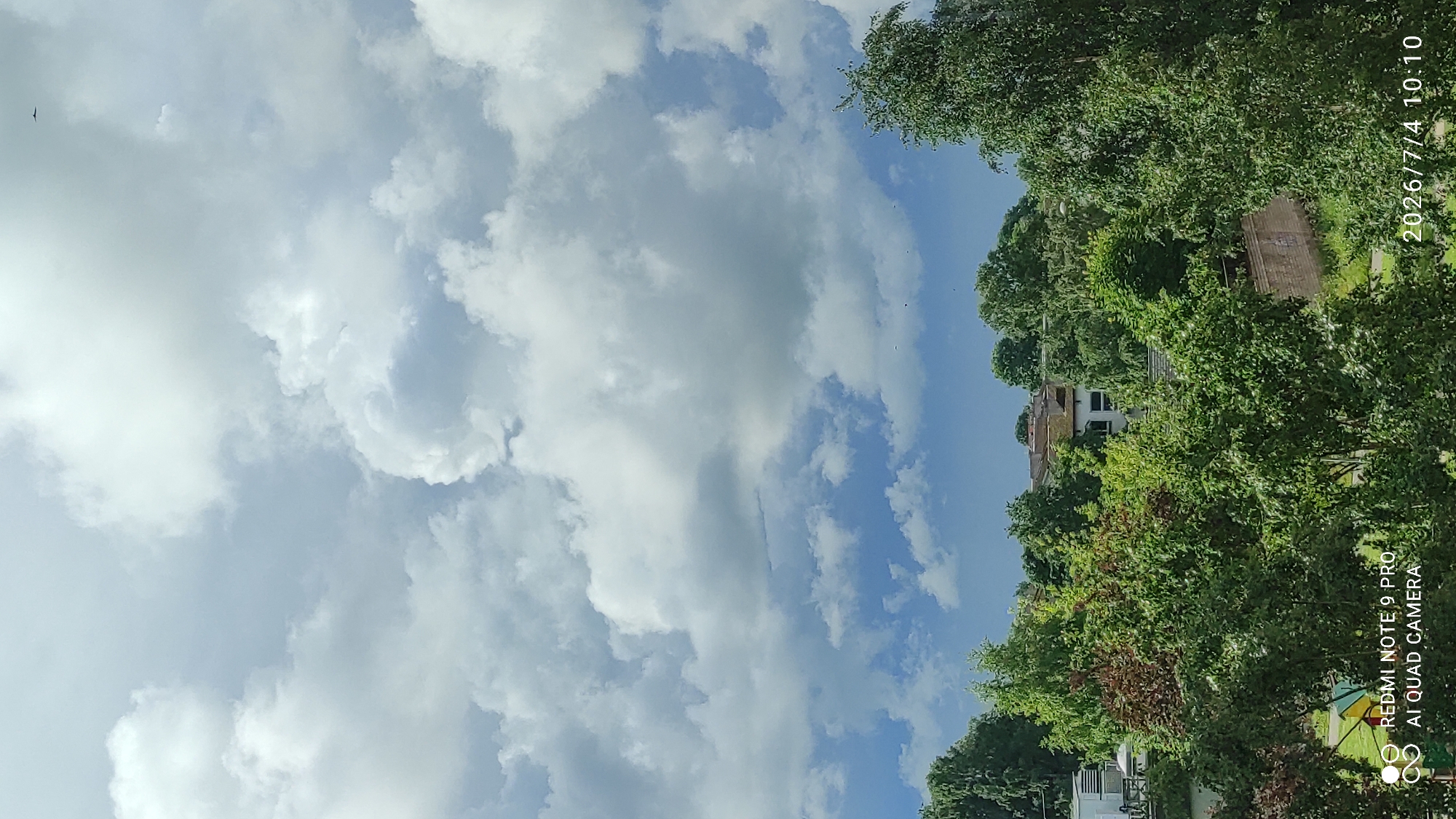 This vertical, full shot captures a view of Maardu, Estonia on July 4, 2026, at 10:10 AM. The sky is predominantly filled with dramatic, large white and grey cumulus clouds against a backdrop of a lighter blue sky. In the far upper right corner of the sky, a small, dark silhouetted object, likely a bird or insect, is visible.
Below the expansive sky, a dense canopy of green trees dominates the midground and foreground. The trees are lush and varied in their foliage, with some exhibiting reddish-brown leaves interspersed among the green. Peeking through the trees in the midground are several buildings, appearing to be residential or commercial structures with muted colored roofs and white walls. A beige brick wall is visible in the lower right midground, partially obscured by trees. In the lower left quadrant of the image, a small section of a playground with brightly colored equipment is visible. The bottom edge of the image contains watermarks indicating "REDMI NOTE 9 PRO" and "AI QUAD CAMERA" on the left, and the date and time "2026/7/4 10:10" on the right. The overall impression is a serene, overcast day in a tree-lined urban or suburban setting.