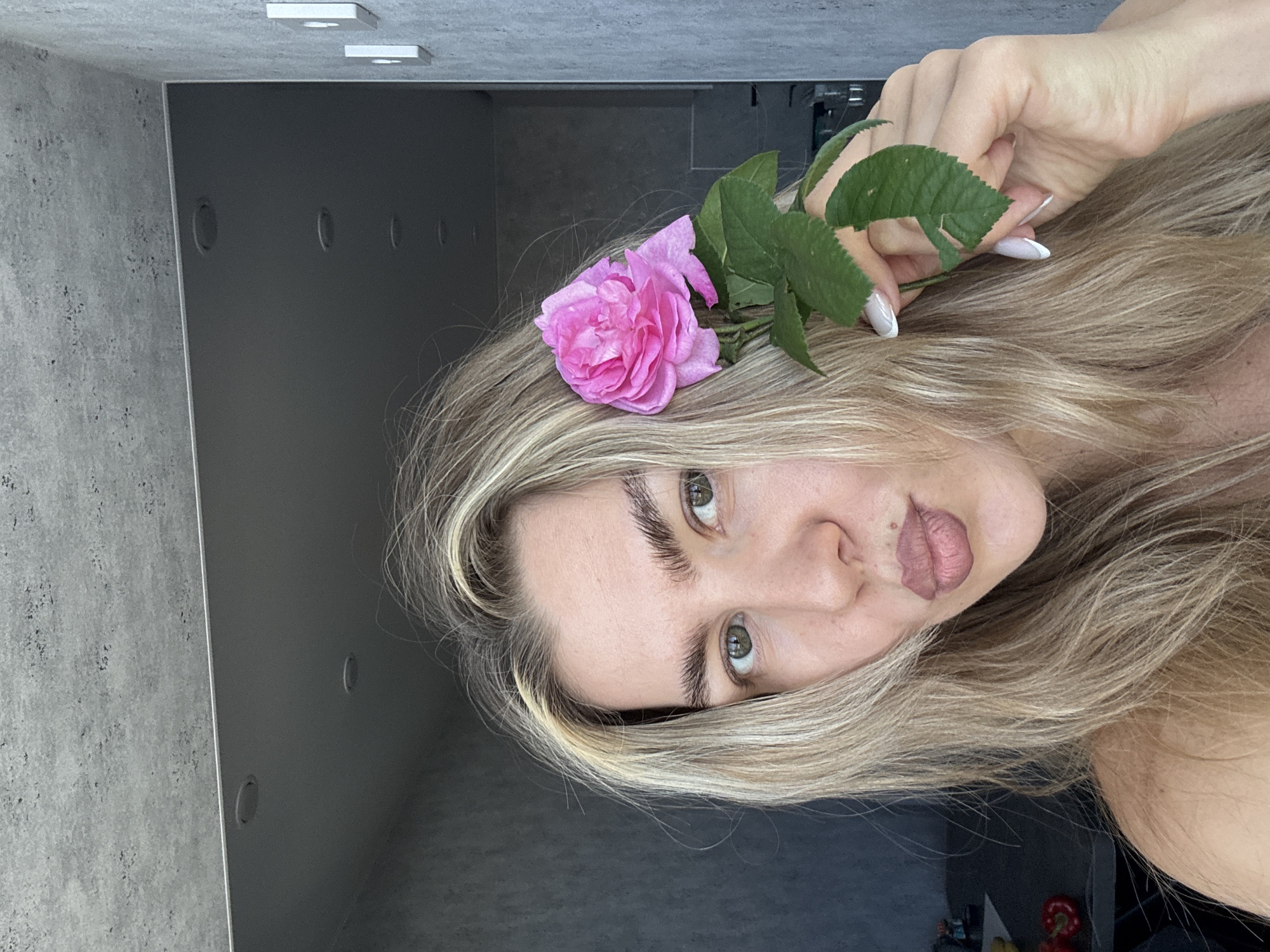 A woman with long, light blonde hair is captured in a close-up, angled shot. She has light-colored eyes (likely green or hazel) and prominent, dark eyebrows. Her lips are full and have a muted pinkish-brown tone, possibly from lipstick or a lip tattoo. A single pink rose with green leaves is held in her hand, which features a French manicure, and is positioned near her head, resting on her hair.
The background consists of a textured, light gray wall, resembling concrete, on the left side. To the right of this wall is a dark gray or black vertical panel, possibly a door or cabinet, with several small, circular indentations or handles visible. Above, a light gray ceiling with two rectangular, white flush-mounted light fixtures is visible. The lighting appears soft and even, suggesting an indoor setting. In the lower right corner, a subtle reflection shows indistinct shapes, including a red object that resembles a bell pepper. The woman's expression is neutral and direct, looking towards the viewer. No text is visible in the image.
