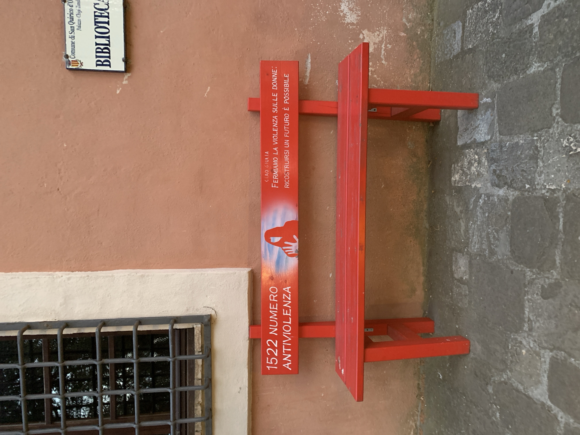 This media file showcases an outdoor scene in San Quirico d'Orcia, Italy, featuring a red metal structure attached to a peach-colored wall. The structure appears to be a display or information stand. On the vertical panel of the stand, white text reads "CIAO GIULIA FERMIAMO LA VIOLENZA SULLE DONNE: RICOSTRUIRSI UN FUTURO È POSSIBILE" and "1522 NUMERO ANTIVIOLENZA". There's also a red silhouette of hands against a sky-blue and white background at the top of the panel. To the left of the stand, a window with a metal grate is visible, set into the wall. Above the window, a white sign reads "Comune di San Quirico d'O Palazzo Chigi-Zond BIBLIOTECA". The background wall transitions to a rough, grey stone masonry. The lighting suggests it is daytime.