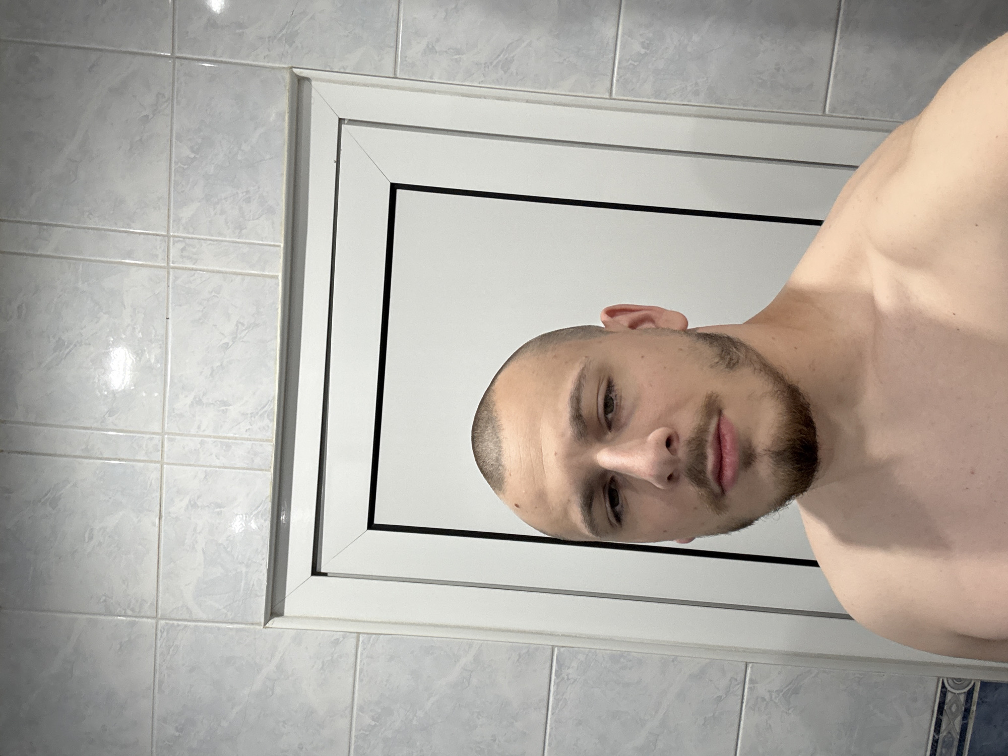 A self-portrait captured indoors shows a young man, shirtless, with his head and shoulders visible. The image is rotated 90 degrees clockwise, making the man appear to be lying on his side or the phone was held in a portrait orientation and then turned. He has a buzz cut and a short beard or stubble, looking directly at the camera with a neutral gaze. His upper body shows muscular definition.
The background consists of light blue and grey marbled wall tiles that have a glossy, reflective surface. Behind the man is a prominent white frame, likely a door or window, featuring a distinct thin black line along its inner edge. A decorative blue and white patterned border tile is partially visible at the bottom left, reinforcing the impression of a bathroom or tiled room.
The activity is clearly a selfie. There are no visible cues for the time of day or external weather. No text is visible within the image. The location context is Stara Zagora, Bulgaria.