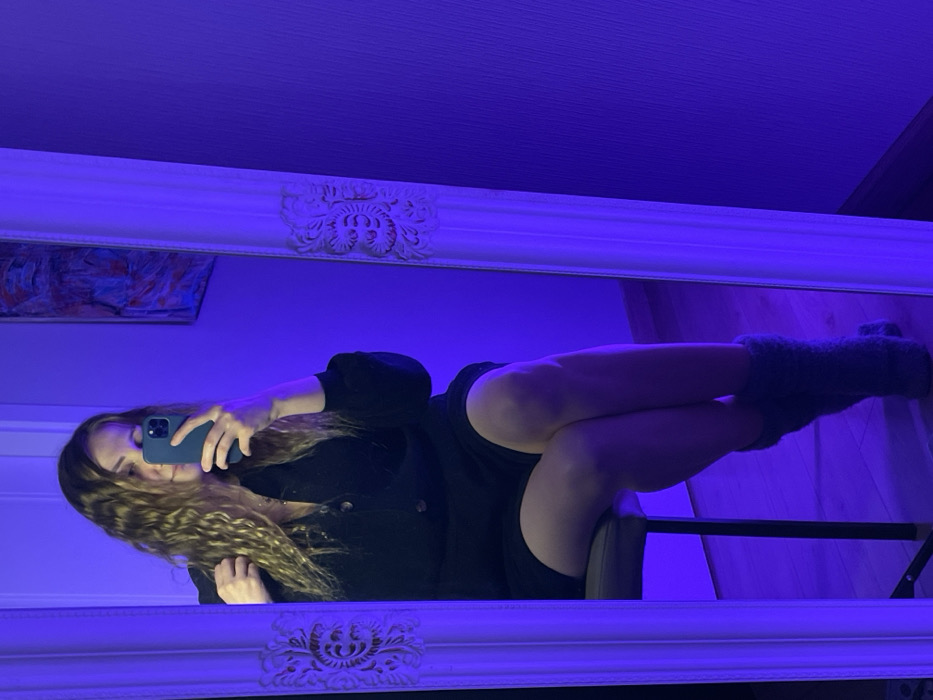 The scene depicts a female individual within a room in the city of Batumi, Georgia, captured in a mirror selfie under strong purple ambient lighting. The subject, possessing long, wavy, blonde hair, is seated on a dark-colored chair with a light-colored cushion, her legs crossed at the knees. She wears a dark, long-sleeved garment with buttons and grey, fuzzy leg warmers. With her right hand, she holds a smartphone encased in a dark-colored cover, pointed towards a large, full-length mirror. This mirror features an ornate white frame with carved floral designs on its upright sides. An abstract artwork displaying red, blue, and white elements is visible on the wall reflected behind the subject. The floor is light-colored, also imbued with the purple light.