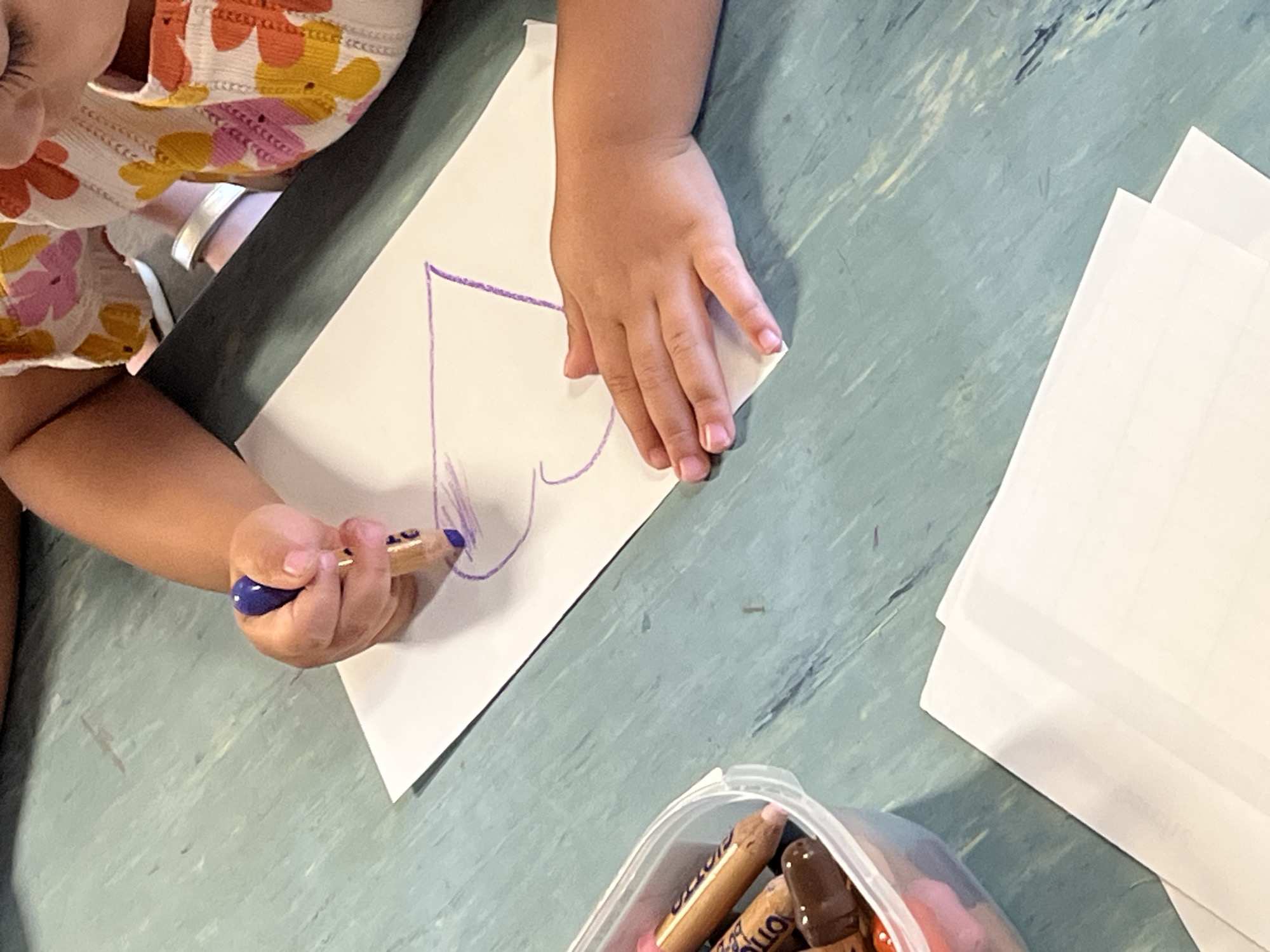 A child is drawing a heart shape on a white piece of paper with a purple crayon. The child is wearing a floral print shirt. A container of crayons is visible in the bottom right corner of the frame, and a stack of white paper is on the right. The picture was likely taken in a home, and the surface appears to be a green countertop. The city of La Fouillouse, France is not discernable in the image.