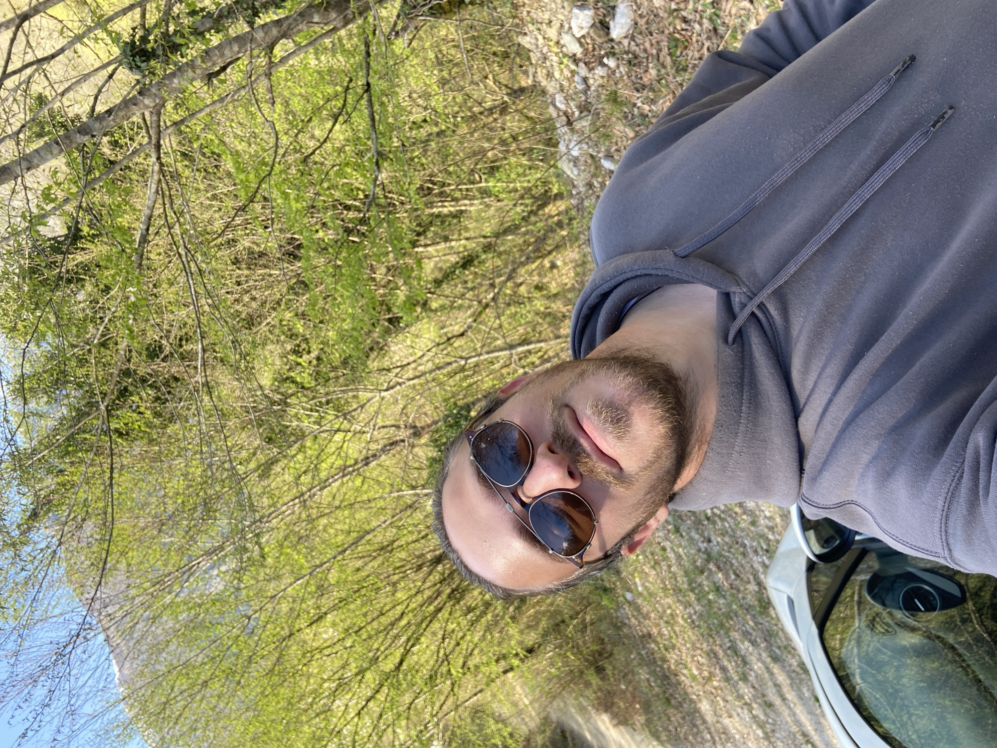 A man with sunglasses and a gray hoodie is standing next to a white car in Mrtvo Duboko, Montenegro. The background is a forest with green trees and bushes. The scene is shot at a slightly rotated angle.