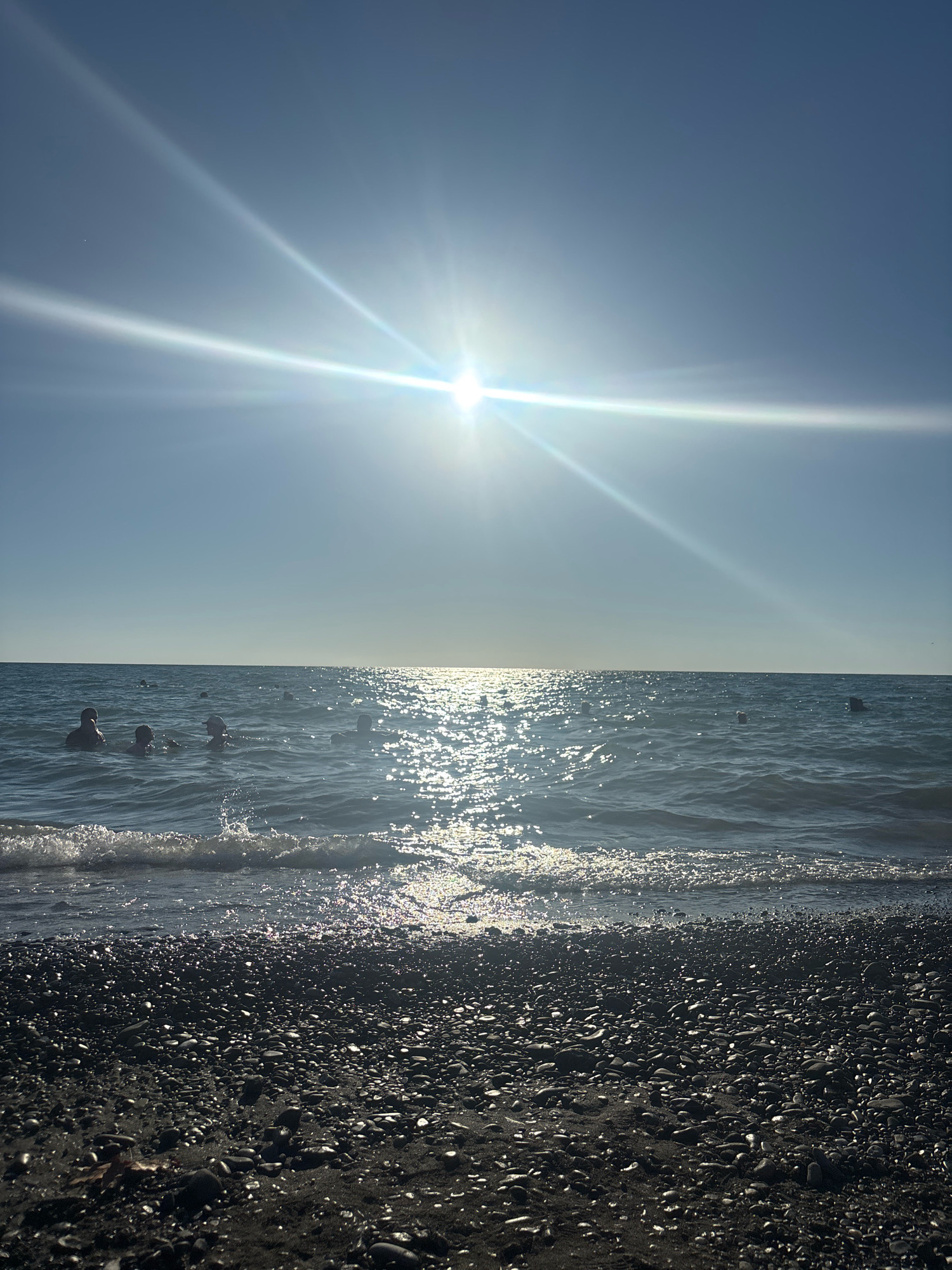 The image depicts a sunny day at a pebble beach on the Black Sea coast in Sochi, Russia. The sun is high in the sky, casting a bright glare and creating a shimmering reflection on the water. Several people are visible in the sea, enjoying the water, with their heads and shoulders above the surface. The foreground of the image is filled with dark, rounded pebbles and sand. The sea is calm with small waves breaking on the shore. The overall impression is one of a pleasant summer day at the beach.