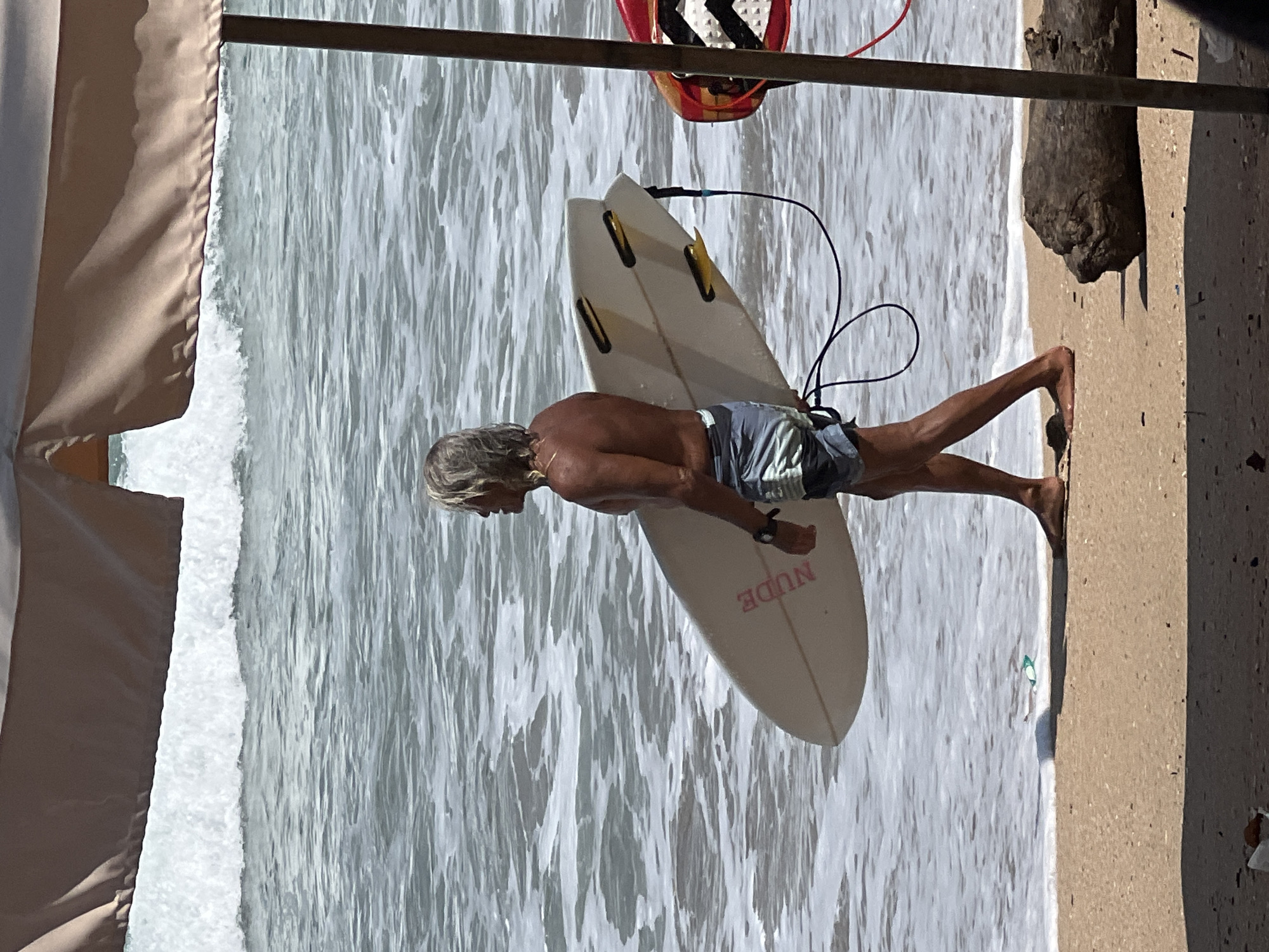 A man with long, grey hair is walking along the shoreline of Kuta Beach in Indonesia, carrying a white surfboard. He is shirtless and wearing striped swim trunks. The ocean has moderate waves with white foam. The beach is sandy, with a piece of driftwood visible in the foreground. A large tan umbrella provides shade in the upper portion of the image.