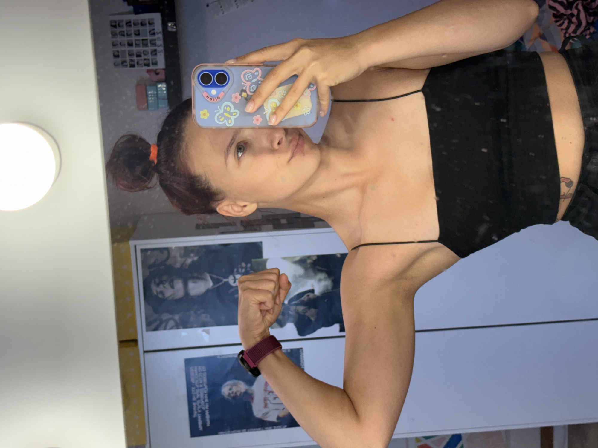 A young woman is captured in a mirror selfie, taken indoors. She has dark hair styled in a bun, secured with an orange tie, and possesses a light complexion. Her gaze is directed towards the smartphone she holds, with a neutral to slightly contemplative expression. She is wearing a black spaghetti strap top.
Her left arm is bent at the elbow, with her hand forming a fist, and a watch featuring a maroon band is visible on her wrist. Her right hand holds a smartphone encased in a light blue protective cover, decorated with various butterflies and flowers. The word "HELLO" is prominently displayed on the phone case, along with the text "H6I10".
The background, reflected in the mirror, reveals elements of a room. A bright, circular wall-mounted light fixture illuminates the scene from the left. Behind the woman's arm, two posters or framed images are visible in the reflection. The upper poster depicts a person with dark hair in a black and white style. Below it, a second poster shows a person with lighter hair, with indistinct, unreadable text accompanying the image. The setting appears to be a casual indoor space, consistent with the context of Прикамский, Russia. The presence of artificial light suggests an indoor time, though specific time of day or weather conditions are not discernible.