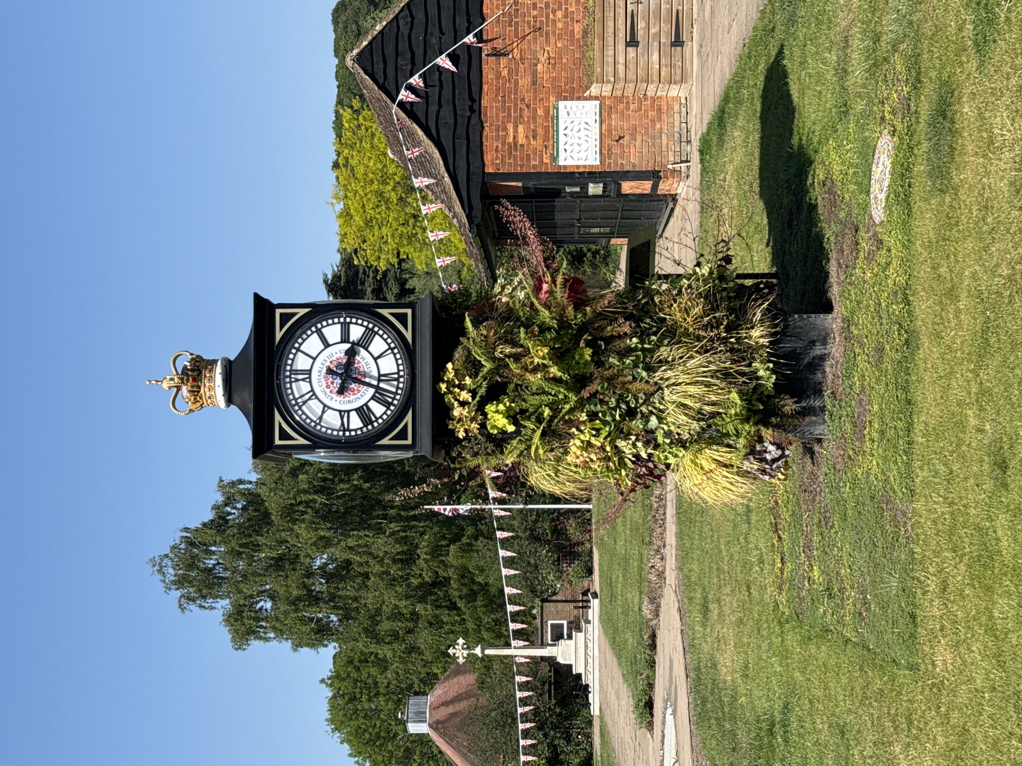 The image shows a black clock with a golden crown on top, situated in a garden-like setting in Amersham, United Kingdom. The clock face is visible with the words "King Charles III Coronation" around it. It is surrounded by plants and greenery. In the background, there is a brick building with black wooden elements. Bunting with British flags is strung around the scene. A stone path leads away from the clock, with a war memorial partially visible further back, decorated with more British flags. The day appears sunny with a clear blue sky.