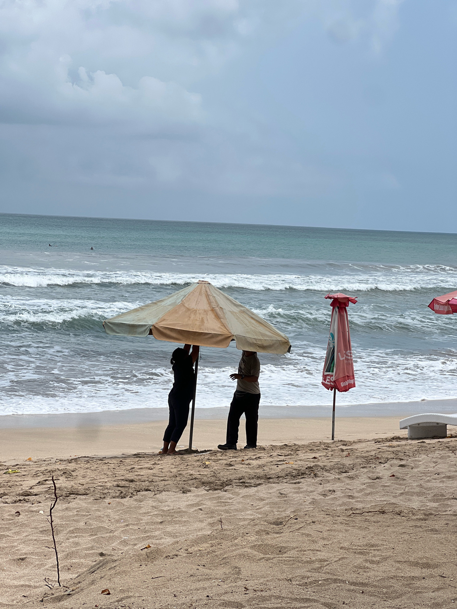 This image captures a beach scene in Kuta, Indonesia, likely on a cloudy day. Two individuals are standing under a large, beige and tan beach umbrella, positioned on the sandy shore. The person on the left, dressed in dark clothing, appears to be holding the umbrella pole. To their right, another person in dark pants and a lighter shirt stands near the umbrella. In the background, the turquoise ocean displays moderate waves with white foam crashing onto the shore. Two small figures, possibly surfers, are visible in the distance on the water. A closed, red and white beach umbrella stands upright to the right of the main umbrella, with some visible text and logos on it. Further to the right, a white lounge chair is partially visible. The sandy foreground shows ripples from the wind or water, with a few scattered small twigs and leaves. The overall atmosphere is serene, with a muted color palette dominated by the sand, sea, and overcast sky.