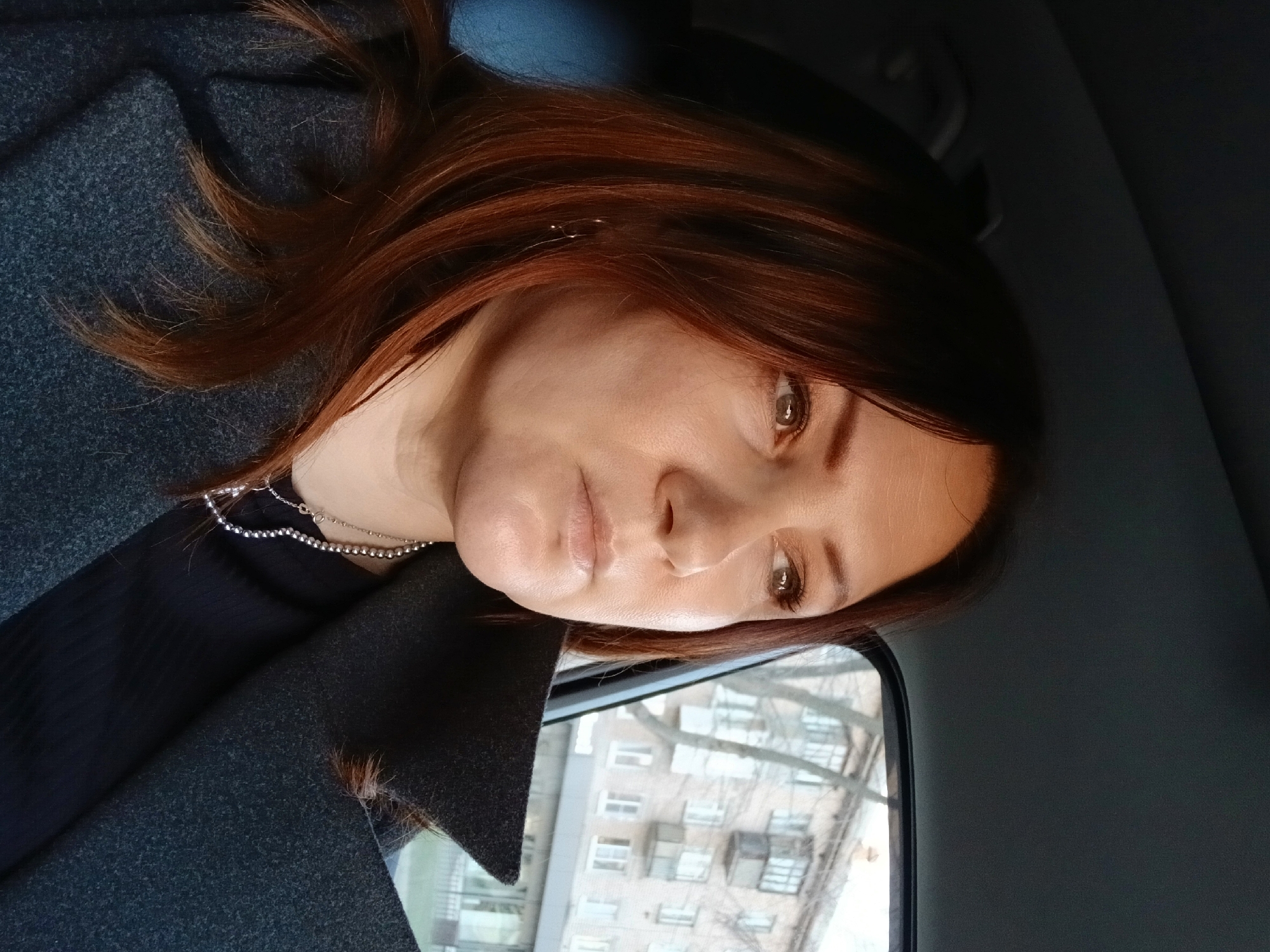 The image features a close-up portrait of a woman inside what appears to be a car or other vehicle. Her head is tilted, and she is looking directly into the frame with a neutral, slightly contemplative expression. She has medium-length, reddish-brown hair that frames her face and flows over her shoulders. Her complexion is fair, and her eyes are light brown. Her lips are slightly parted.
She is dressed in a dark grey or charcoal-colored coat with a visible collar, likely made of a wool or felt-like fabric. Underneath, a black top is visible. She wears two silver necklaces: one is a strand of small silver beads, and the other is a finer, delicate silver chain.
Through the window on the right side of the image, parts of a light-colored, multi-story building with multiple windows and balconies are visible. Bare tree branches are silhouetted against the building, suggesting late autumn or winter. This background indicates an urban or suburban setting.
The lighting is soft and even, suggesting natural light on an overcast day or indirect sunlight. The overall mood is calm. There is no visible text in the image.