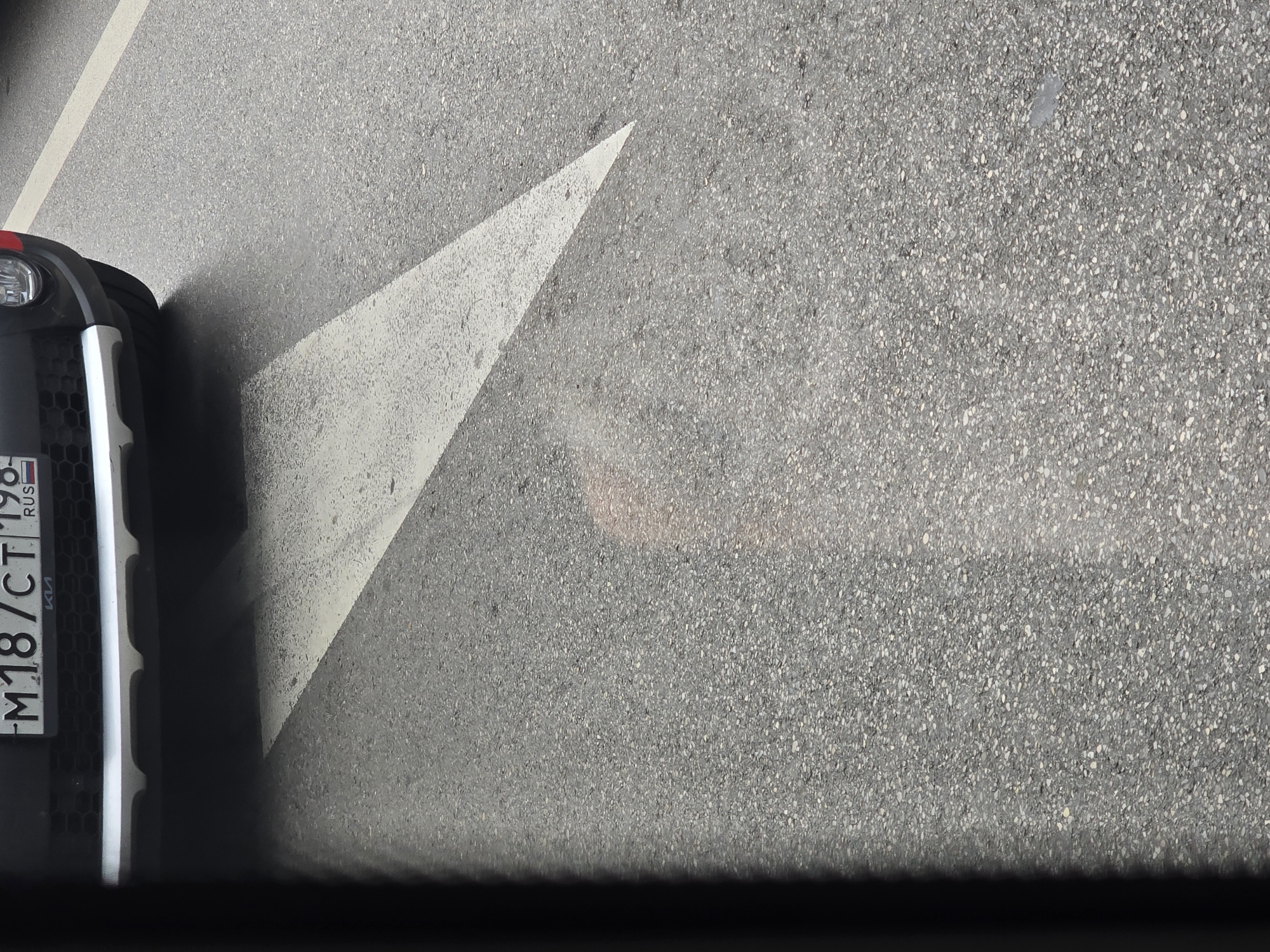 A close-up, downward-angled shot shows the front bumper and license plate of a dark-colored car. The license plate reads "M18/CT 198 RUS". The car is parked on asphalt, on what appears to be a street. A white arrow, painted on the asphalt, points to the right and slightly forward, indicating a direction on the road. A faint shadow or reflection is visible on the asphalt, possibly of the car or a surrounding object. The background consists of more asphalt. The location is not discernible from the image itself, and therefore, cannot be specified as Tbilisi, Georgia.