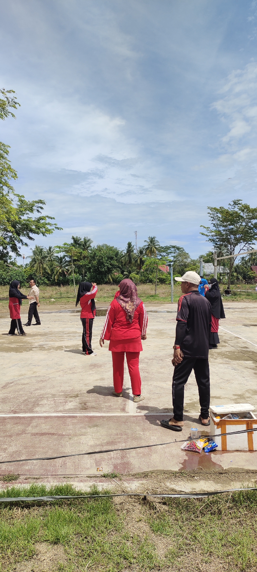 A group of six people, five women and one man, are gathered on a concrete court, likely for recreation or exercise. Four of the women are wearing hijabs, and all are dressed in athletic attire, primarily red and black. One woman in the foreground, facing away from the camera, wears a red tracksuit with white stripes and a red hijab. To her left, another woman in a red tracksuit and black hijab is positioned as if about to engage in activity. Further left, two women in red and black athletic wear and hijabs appear to be interacting. The lone man, also facing away from the camera, wears a black long-sleeved shirt, black pants, and a light-colored cap. He stands to the right of the women. In the background, a sparse, dry grassy area transitions to a line of trees, including palm trees, and houses, suggesting a semi-rural location. A utility pole is visible in the distance. The sky is partly cloudy with a light blue hue. The scene is likely set in Peurupok, Indonesia, given the attire of the individuals and the tropical vegetation. A white cooler and some bags of snacks are present on the edge of the court in the lower right portion of the frame.
