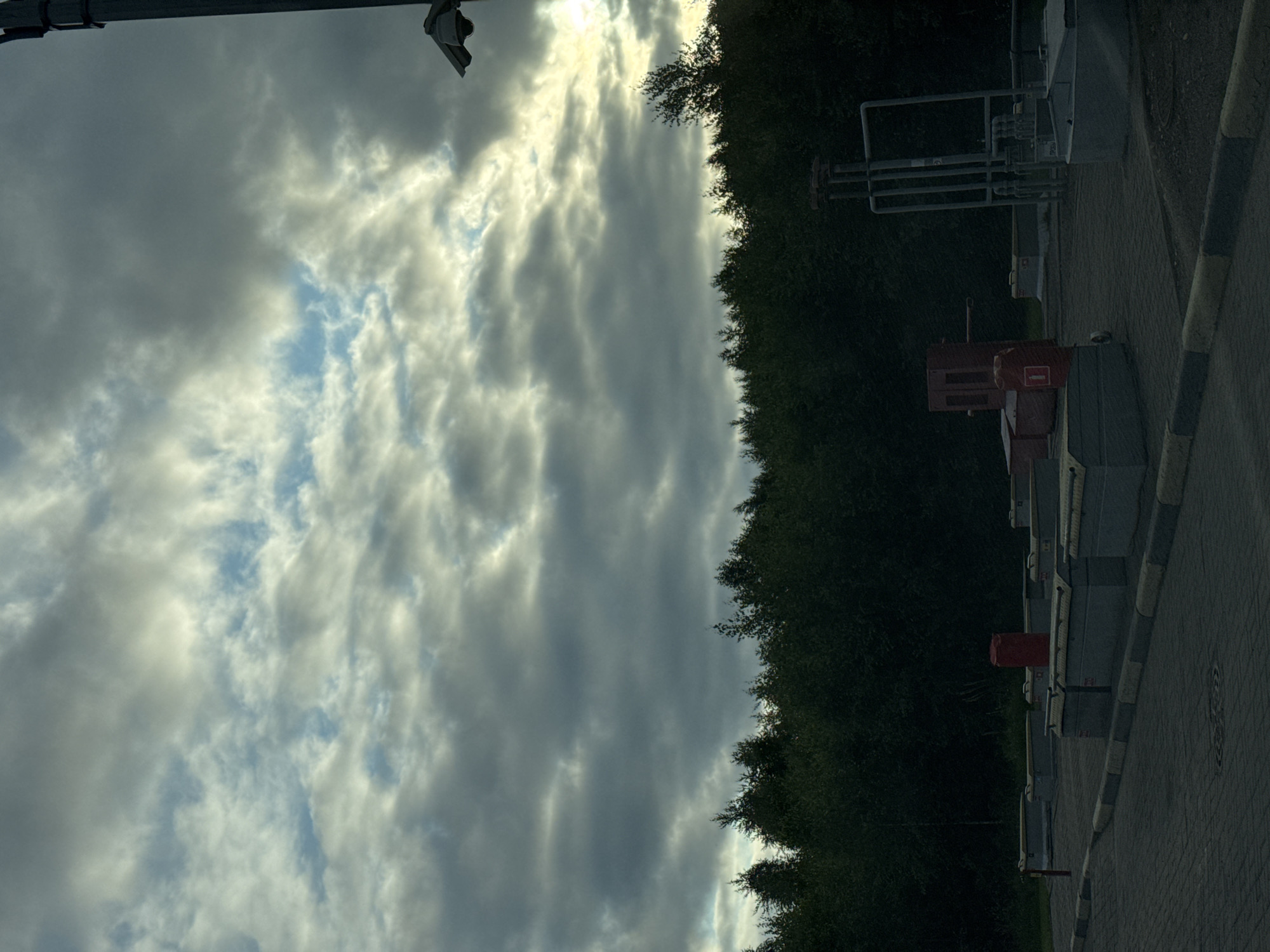This image shows an outdoor industrial area in Kolyasovo, Russia. In the foreground, there is a paved area with several concrete structures, some of which appear to be containment units. A red cylinder with a white fire extinguisher symbol is visible. Behind these, a dense line of dark green trees forms the background. Above the trees, the sky is filled with a dramatic display of grey and white clouds, with patches of blue sky peeking through. The lighting suggests it is either early morning or late evening. A security camera is mounted on a pole on the right side of the image, overlooking the area.