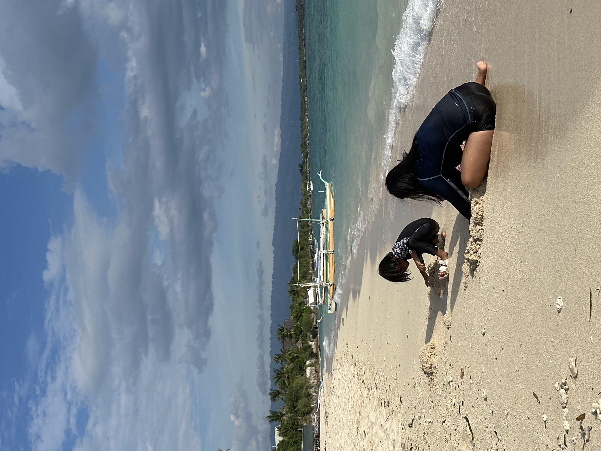 The image depicts a beach scene in Saavedra, Philippines. Two individuals, likely children, are engaged in playing on the sandy shore. One person, wearing a black rash guard and black shorts, is crouched down and actively playing in the sand. The other person, also in a black rash guard and black shorts, is positioned slightly further away and appears to be looking at something in the sand. The environment includes a clear blue ocean with gentle waves lapping at the shore, a sandy beach with scattered shells and debris, and lush green trees lining the coast in the distance. Several traditional Filipino outrigger boats are visible in the shallow water. The sky is a mix of blue and grey clouds, suggesting a partly cloudy day. The overall impression is one of a tranquil beach setting with recreational activity.